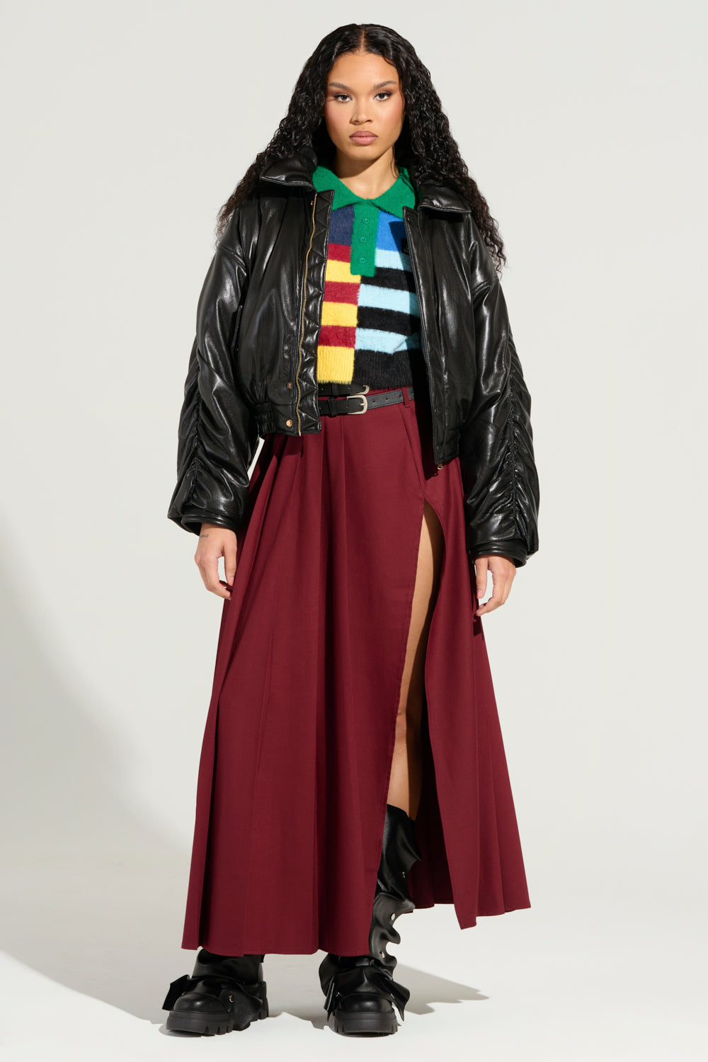 A person with long hair wears a colorful striped sweater under the TOKYO DRIFTING FAUX LEATHER BOMBER, paired with a long maroon skirt featuring a high slit. They complete the look with black boots, exuding an effortless Tokyo drifting vibe against a plain background.