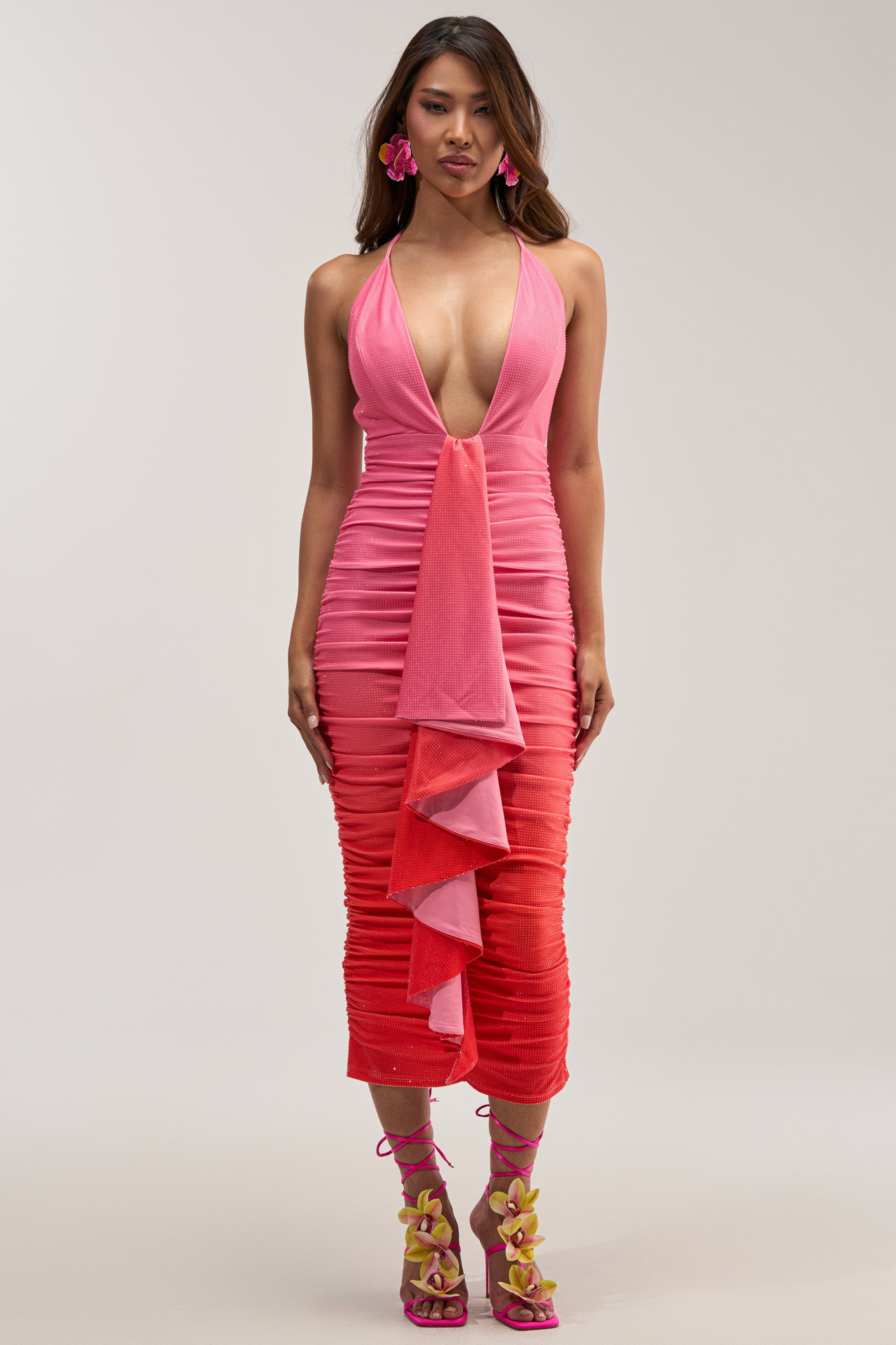 Woman in the AMAYA RHINESTONE MIDI DRESS—a sleeveless, deep V-neck pink and red ombre dress with front ruffle detail—paired with matching pink earrings and strappy heels featuring yellow flower accents, standing against a plain light background.