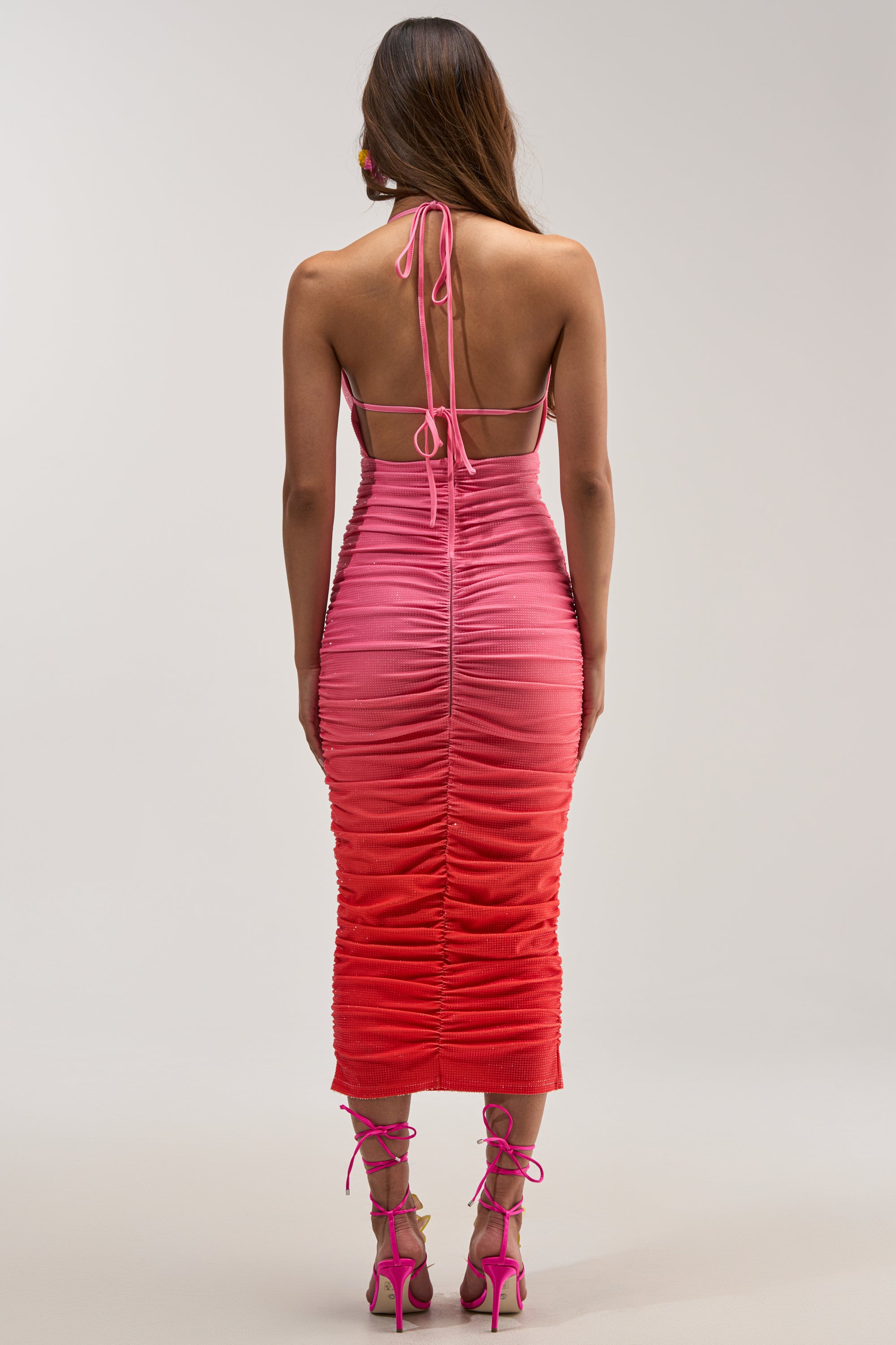 A woman stands facing away, wearing the AMAYA RHINESTONE MIDI DRESS—a vibrant pink and red ruched halter summer party dress with an open back and tie straps—paired with matching high-heeled sandals.