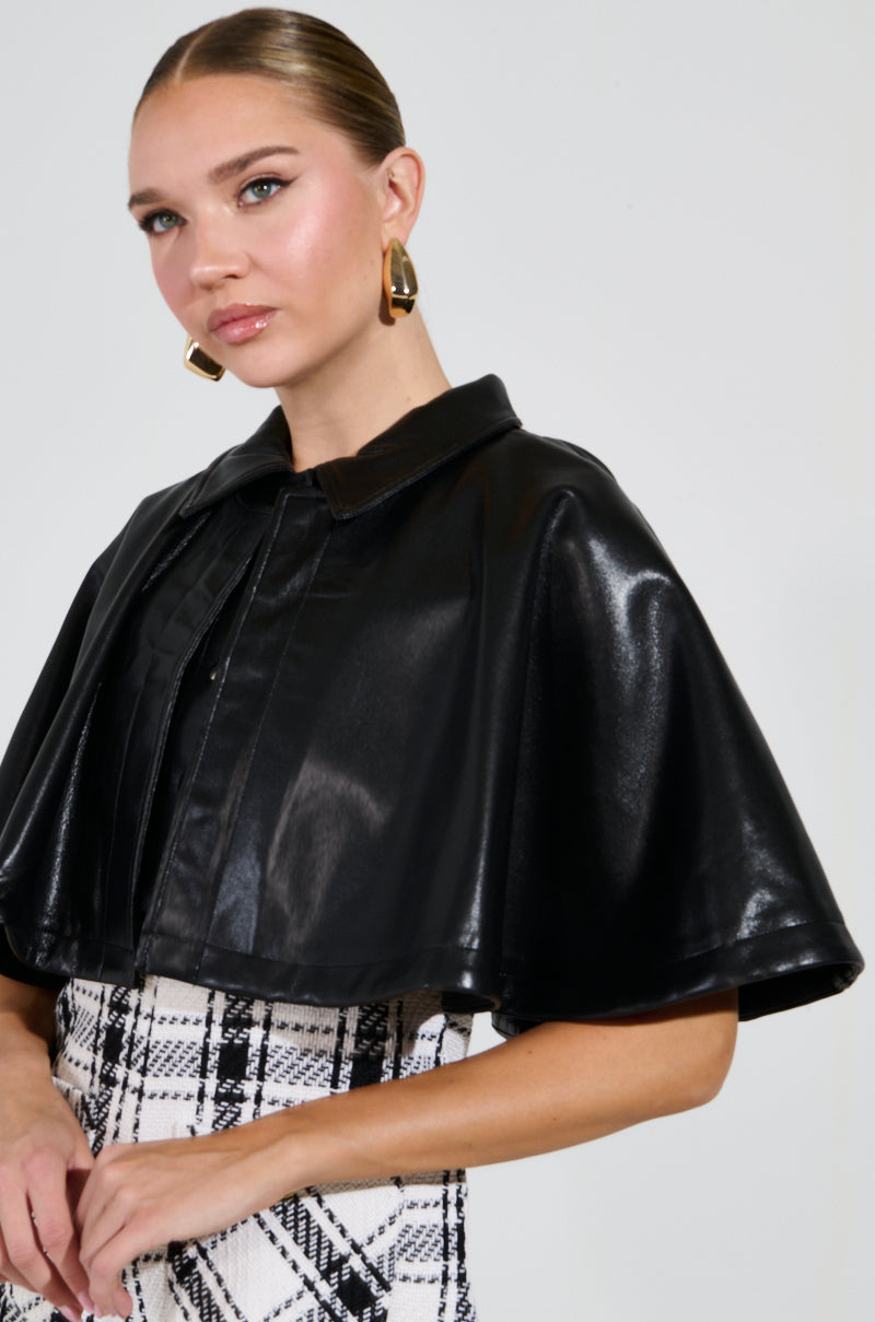 A woman with tied-back hair is wearing a shiny black LOUIE DOUBLE LAYER CAPE over a black and white plaid outfit. She accessorizes with large geometric earrings featuring a dual-layer design and poses against a plain, light background.