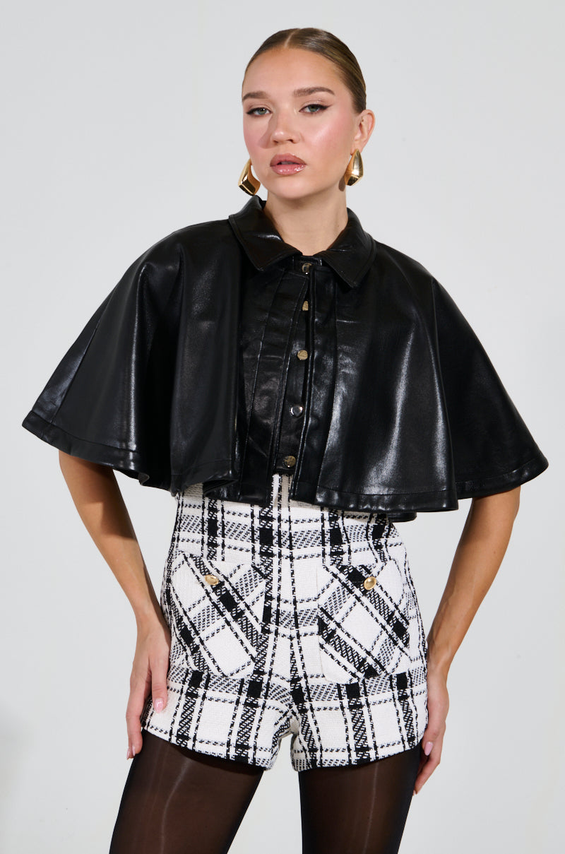 Wearing the LOUIE DOUBLE LAYER CAPE over white and black plaid shorts, a person confidently pairs their look with gold hoop earrings against a plain background.