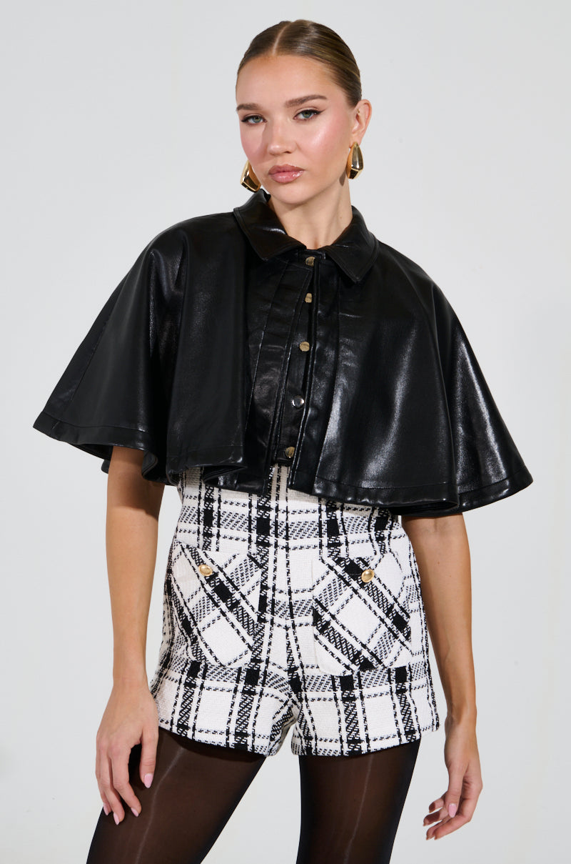 A person exudes confidence wearing the LOUIE DOUBLE LAYER CAPE, paired with black-and-white plaid shorts. Their hair is styled back and complemented by gold hoop earrings, all set against a neutral background.