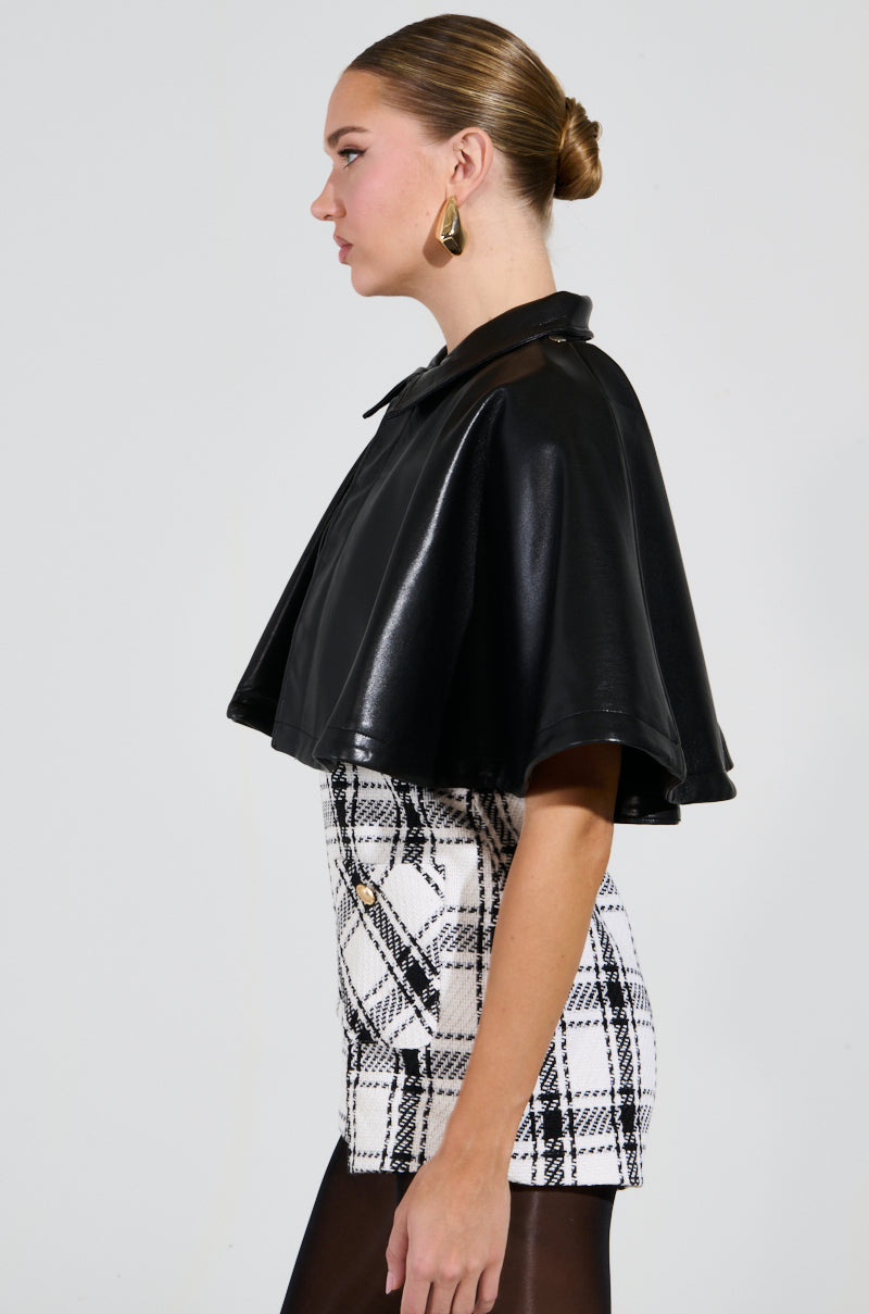 A woman with styled hair is wearing a LOUIE DOUBLE LAYER CAPE in black faux leather, paired with a black-and-white plaid dress. She accessorizes with gold hoop earrings and poses in profile against a plain background, highlighting the dual-layer design of her outfit.