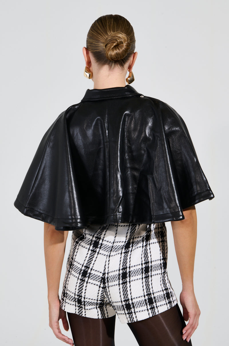 A person with blonde hair in a bun is seen from behind, wearing the LOUIE DOUBLE LAYER CAPE in black faux leather, along with checkered shorts and black tights. The look hints at a dual-layer design against the plain, light background.