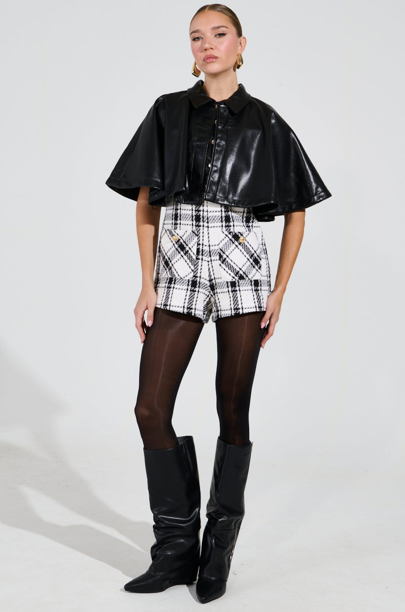 A person is elegantly posed against a plain white background, wearing the striking LOUIE DOUBLE LAYER CAPE in black faux leather over a collared shirt. This ensemble is paired with black and white plaid shorts, sheer tights, and black knee-high boots.