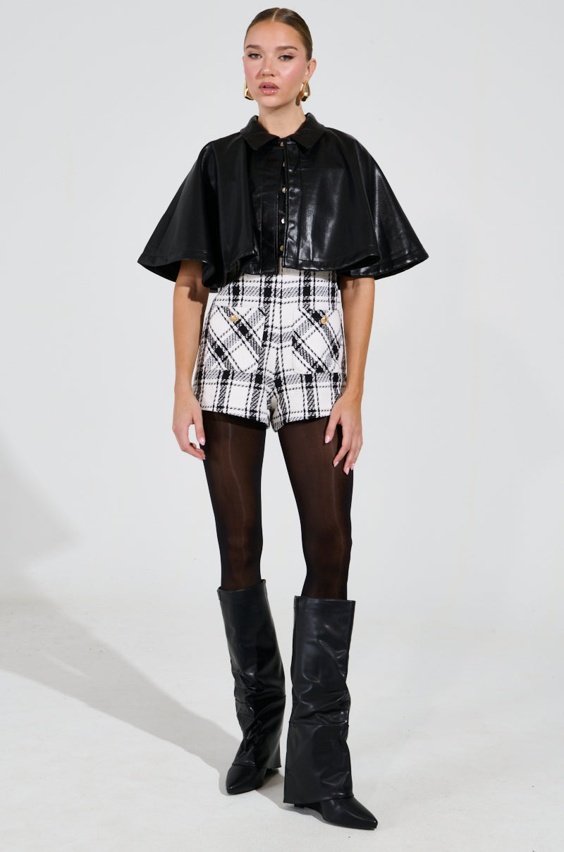 A person poses confidently against a plain background, wearing the LOUIE DOUBLE LAYER CAPE, paired with plaid shorts, black tights, and knee-high boots. They sport large hoop earrings and have their hair neatly pulled back.