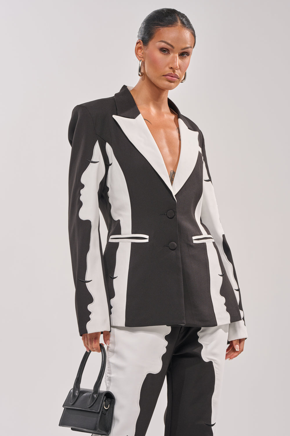 A woman with her hair pulled back stands confidently against a plain background, wearing the POV GRAPHIC BLAZER featuring a black and white abstract face design and carrying a small black handbag. 0