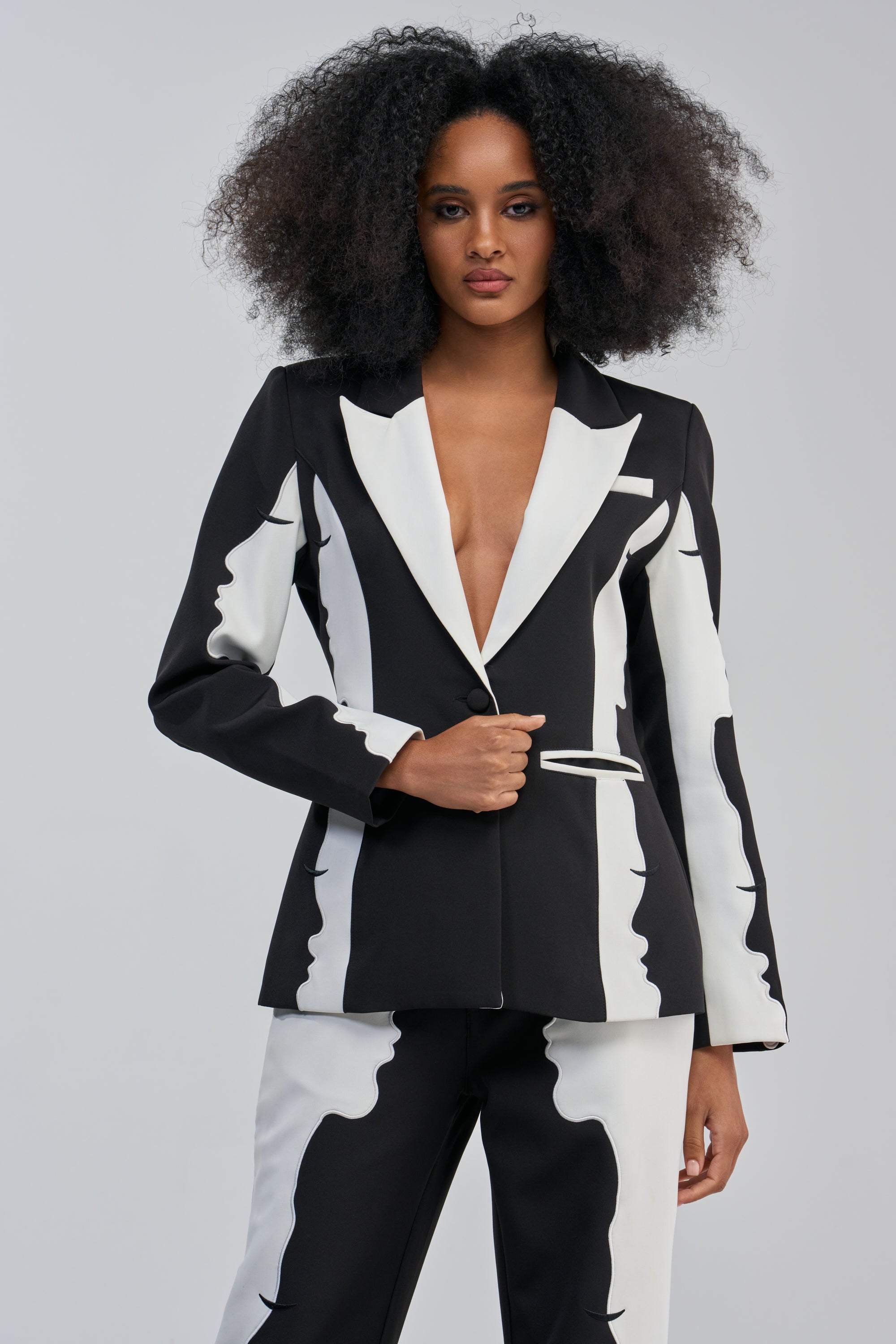 A woman with voluminous curly hair models the POV GRAPHIC BLAZER, featuring a striking black and white abstract color block design and a deep V-neck, paired with matching pants and posed against a plain light gray background.