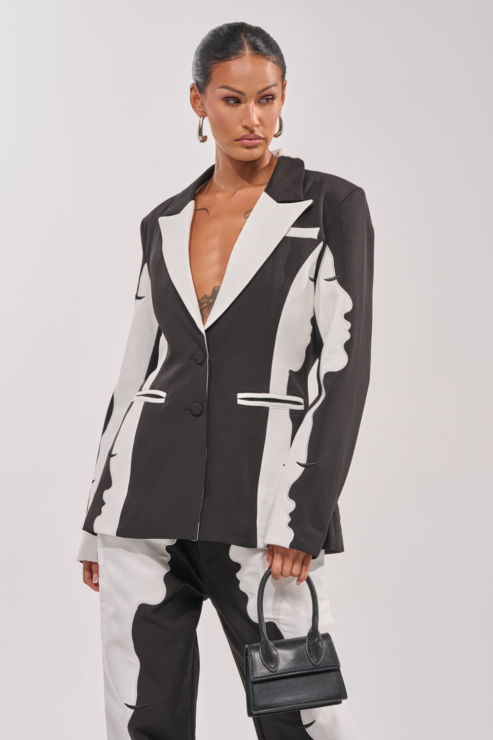 A woman poses confidently against a plain light background in the POV GRAPHIC BLAZER, paired with a small black handbag and hoop earrings. Her slicked-back hair completes this effortless take on power dressing. 1