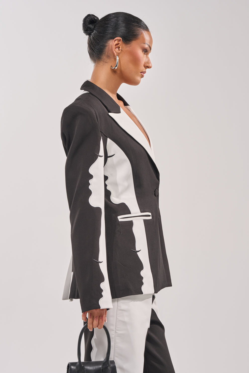 A woman with slicked-back hair exudes confidence in the POV GRAPHIC BLAZER, a black and white piece featuring profile silhouettes. She accessorizes with statement earrings and a small black handbag, standing poised against a plain backdrop. 3