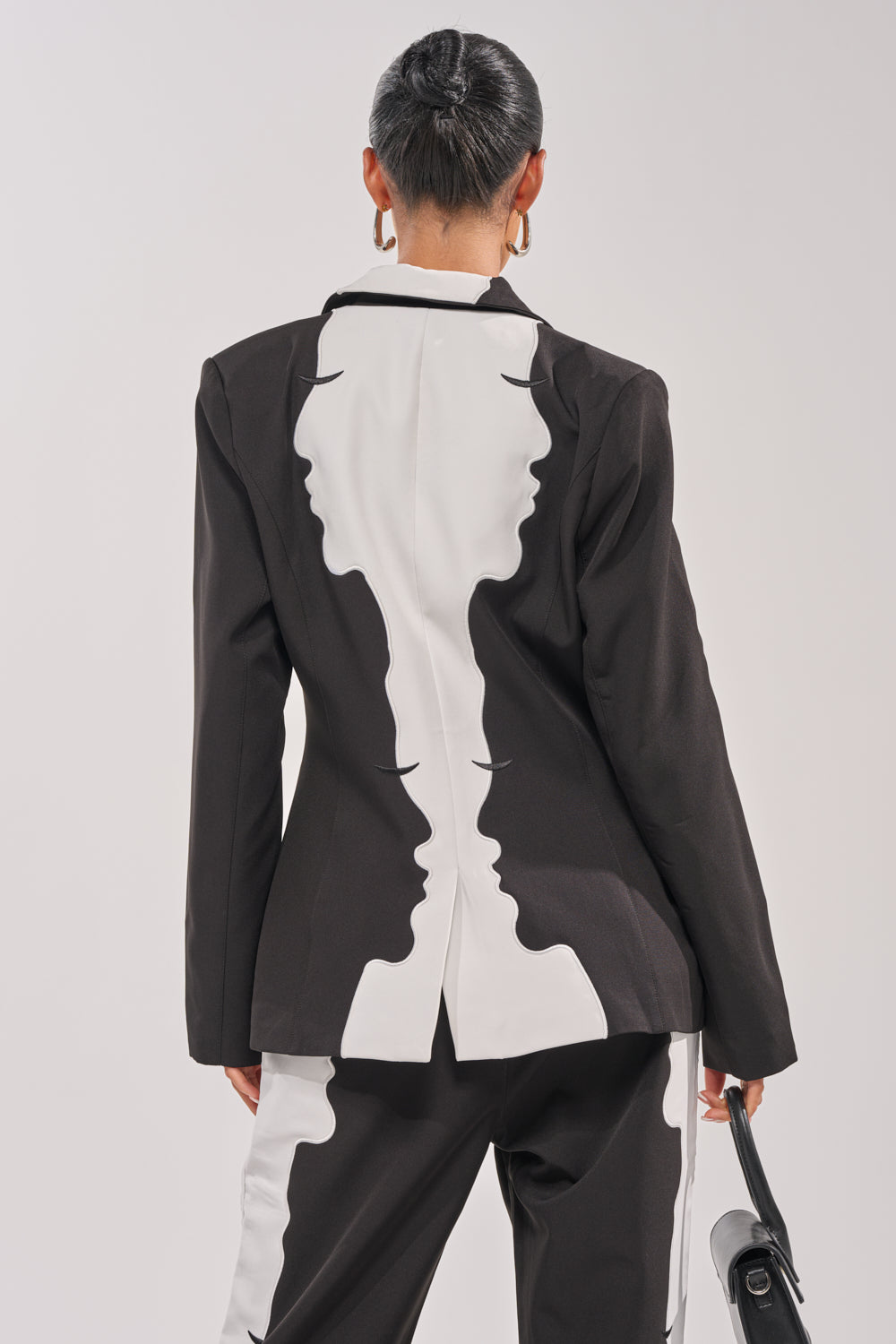 A person with hair in a neat bun wears the POV GRAPHIC BLAZER featuring abstract face profiles on the back and matching pants, paired with a black handbag, photographed from behind against a plain background. 4