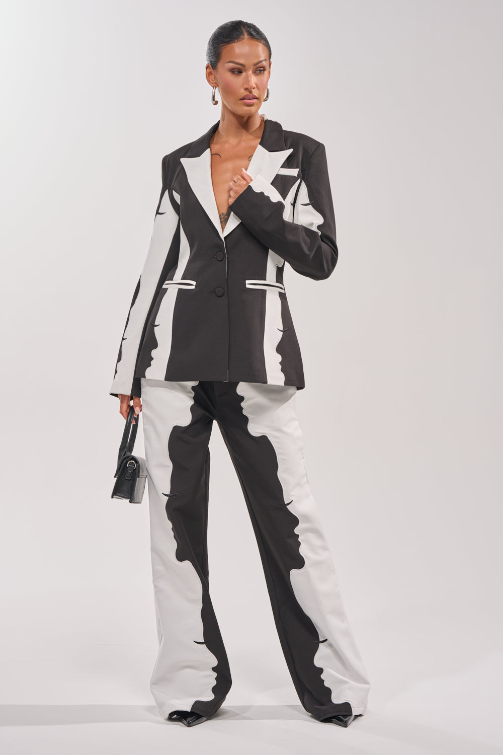 A woman poses confidently in the POV GRAPHIC TROUSER—a high-rise, black and white wide-leg suit trouser with an abstract pattern—paired with a matching blazer and small black handbag against a plain light background. 0