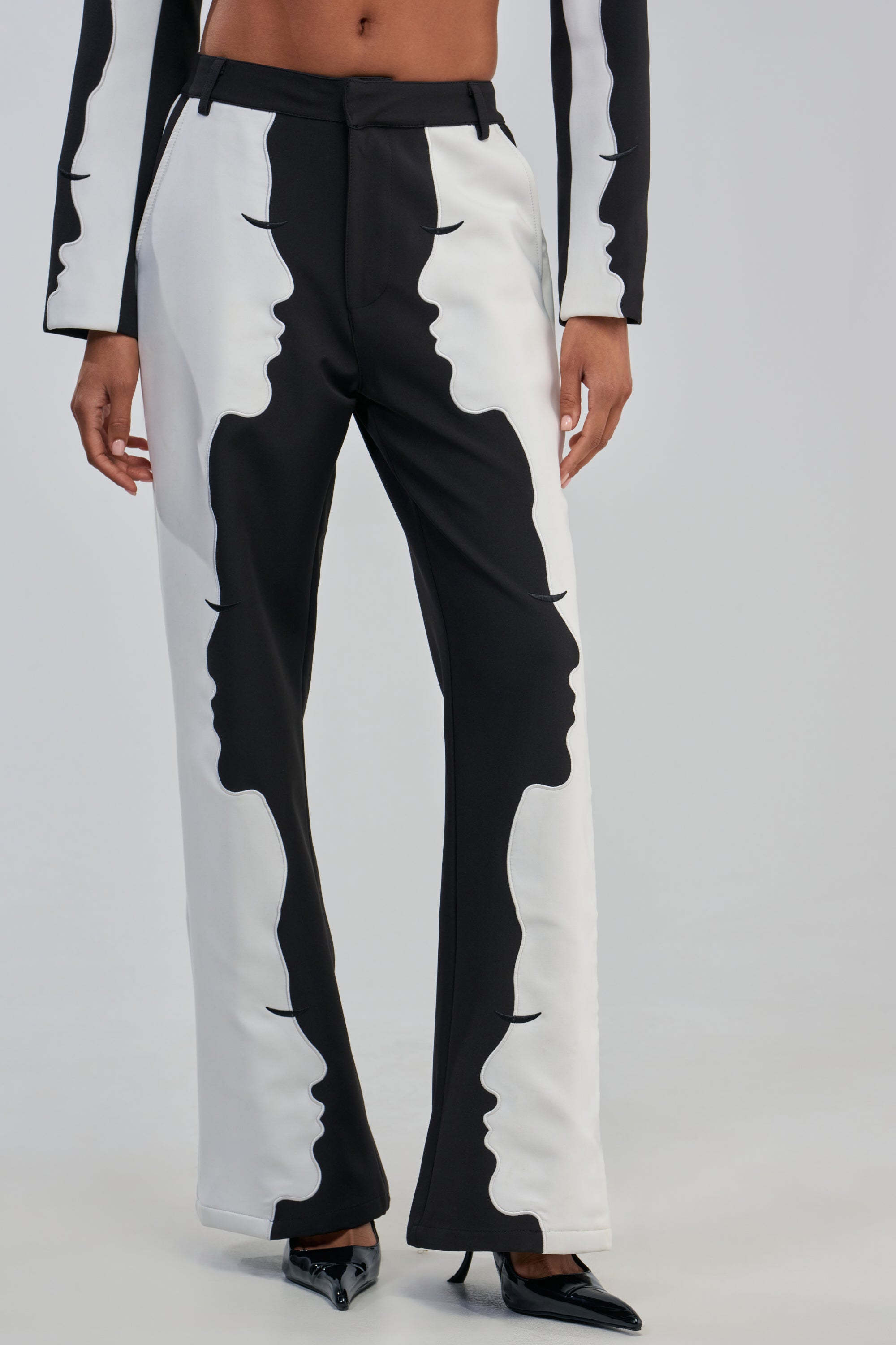 Wearing the POV GRAPHIC TROUSER, which features high-rise wide-leg tailoring with abstract face profile designs along the legs, this individual pairs them with black pointed shoes and a matching jacket, standing against a plain background.