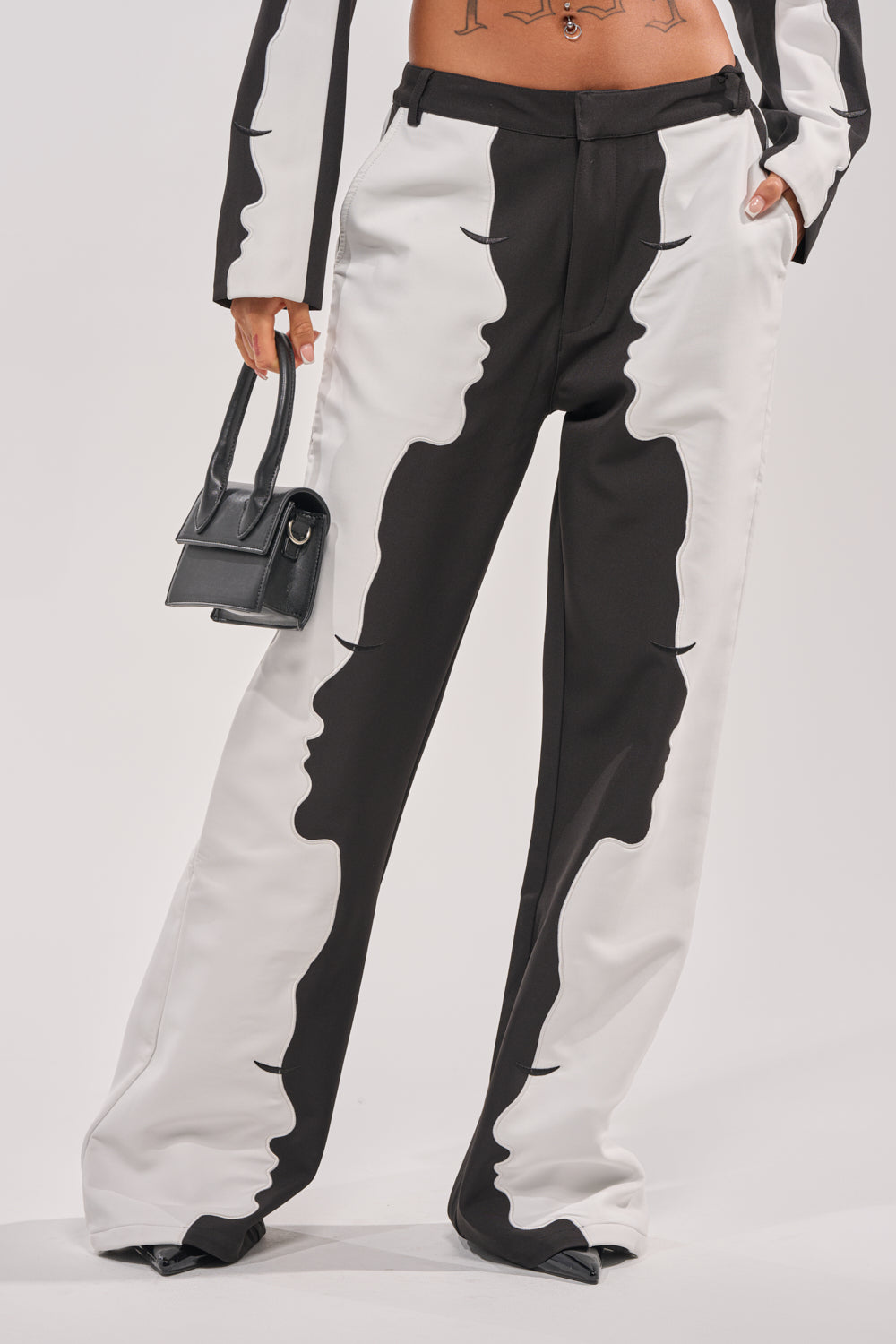 Wearing the POV GRAPHIC TROUSER—black and white wide-leg pants with mirrored face profiles—a person reveals a tattooed midriff above the waistband and carries a small black handbag. 1
