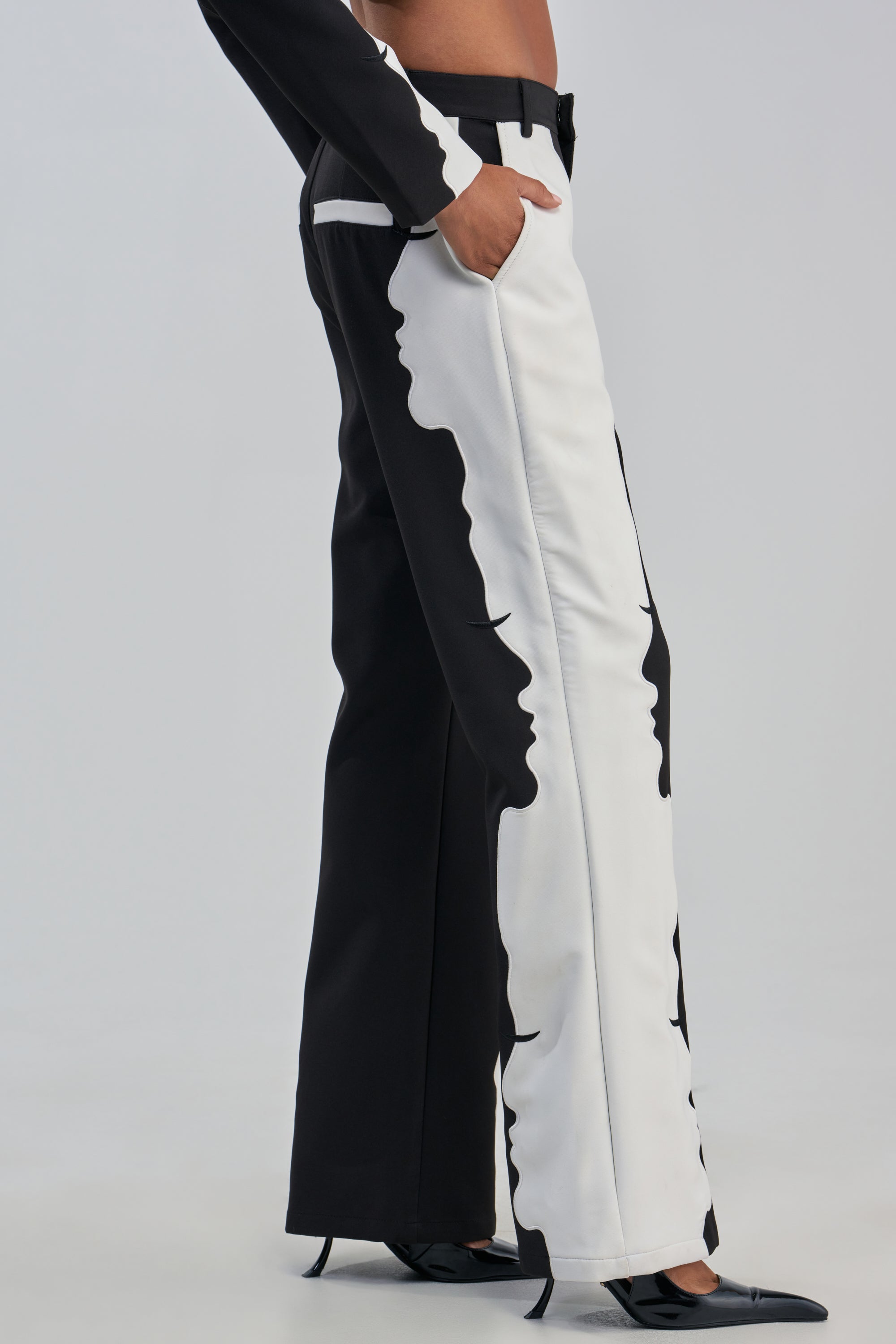 A person wearing the POV GRAPHIC TROUSER, featuring a high-rise wide-leg silhouette with a face graphic design along the side, posing with one hand in a pocket and styled with black pointed-toe shoes.