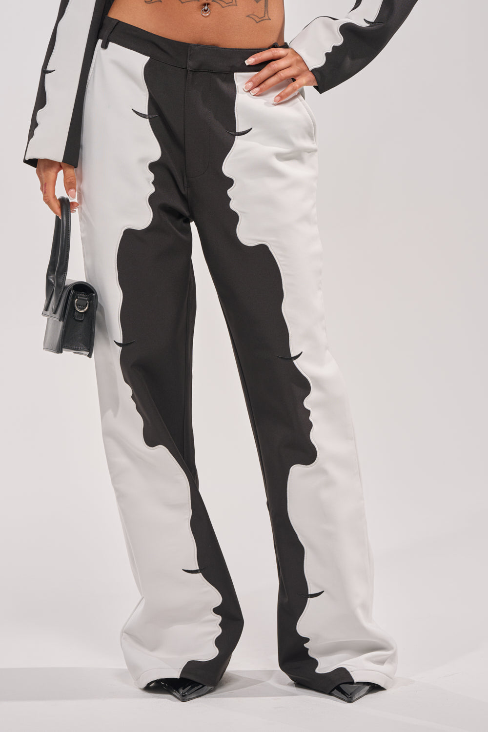 A person wears POV GRAPHIC TROUSER—black and white trousers with mirrored face silhouettes along the legs and a high-rise fit—while holding a small black handbag. The photo is cropped from mid-torso down. 2