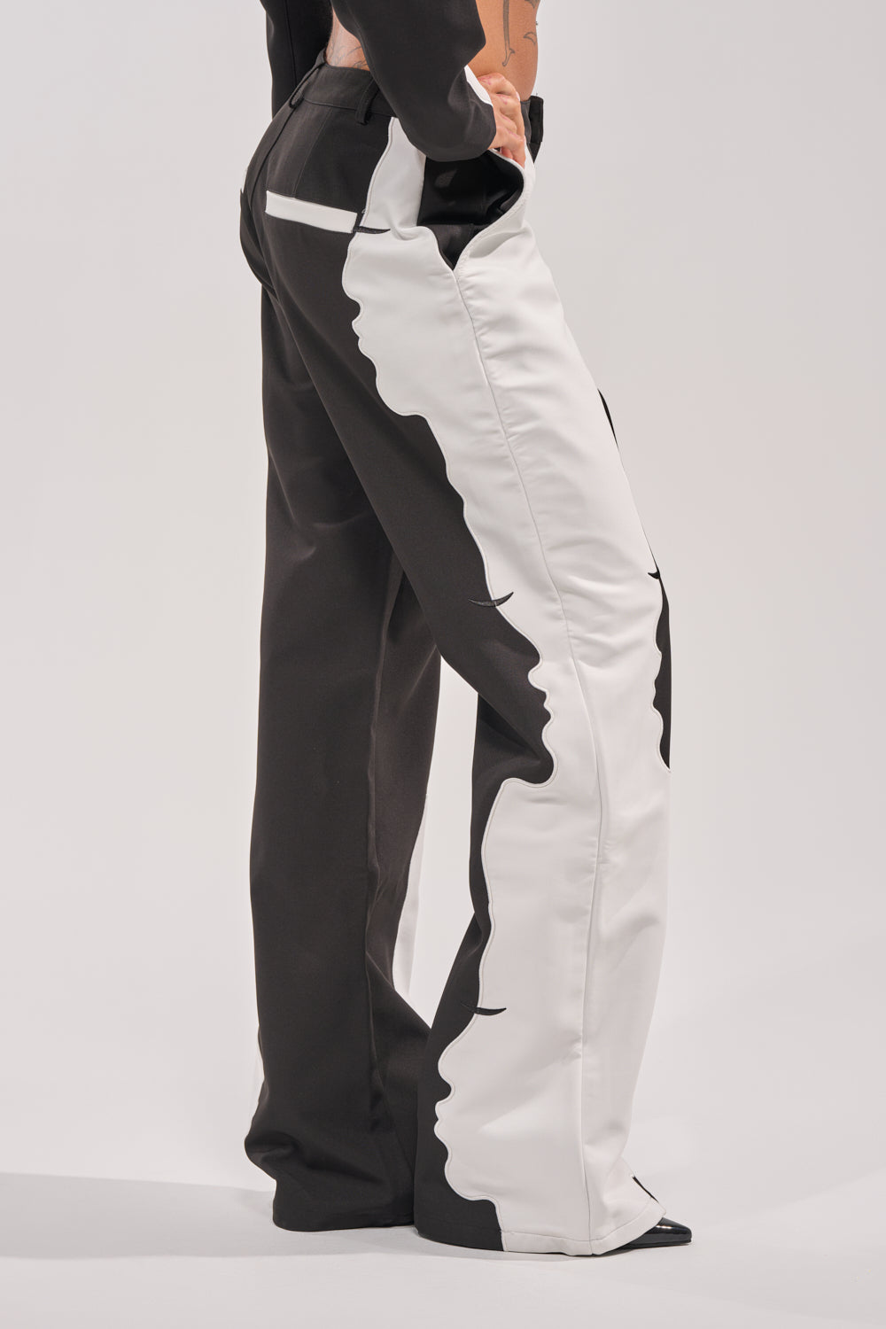 Wearing the POV GRAPHIC TROUSER—high-rise, black and white wide-leg pants featuring bold abstract face profiles along the side seams—a person stands against a plain background. 3