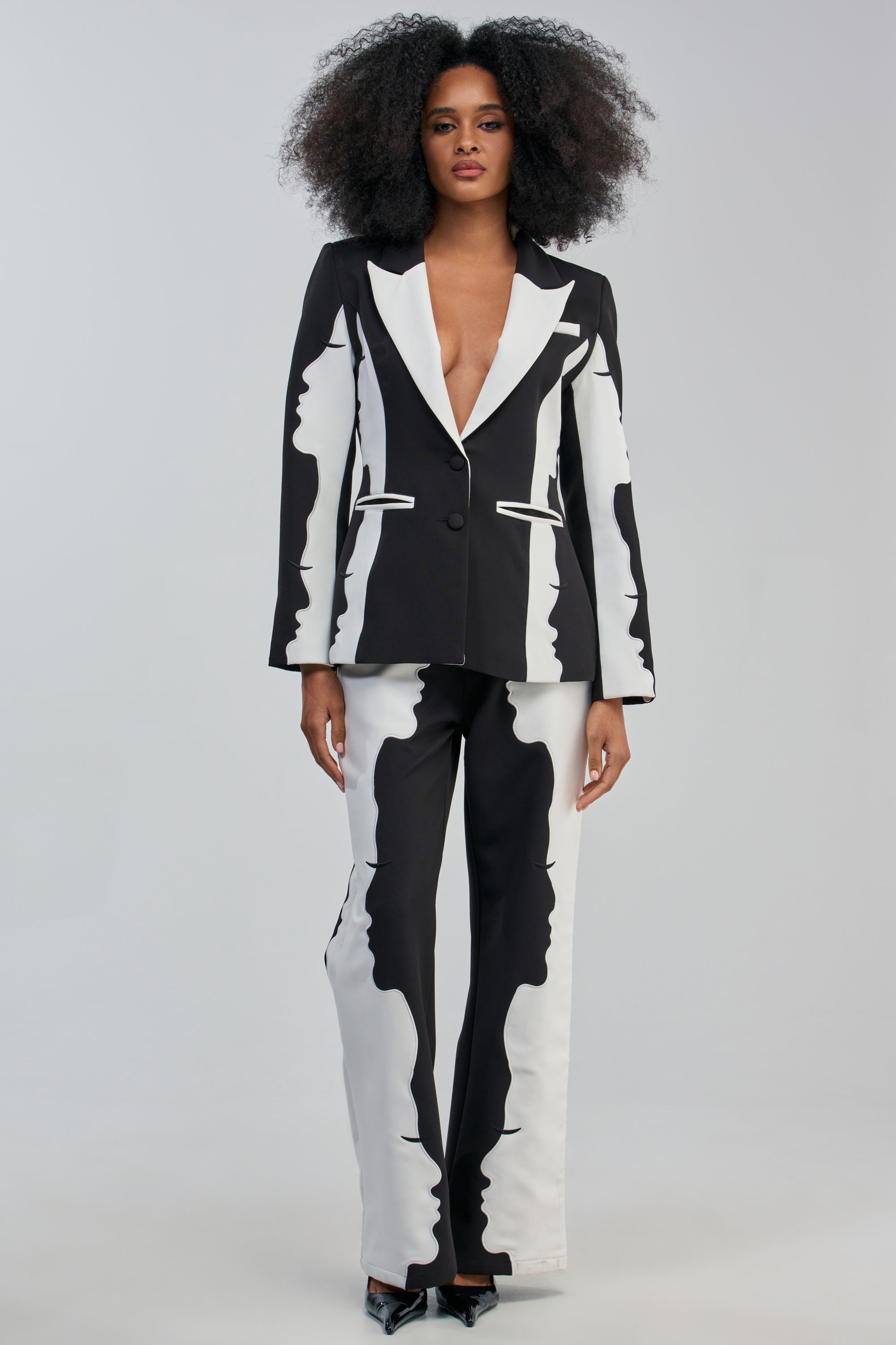 A woman with voluminous curly hair wears a black and white suit with abstract patterns, featuring the POV GRAPHIC TROUSER paired with a deep-neckline blazer, all set against a plain light background for a bold and sophisticated look.