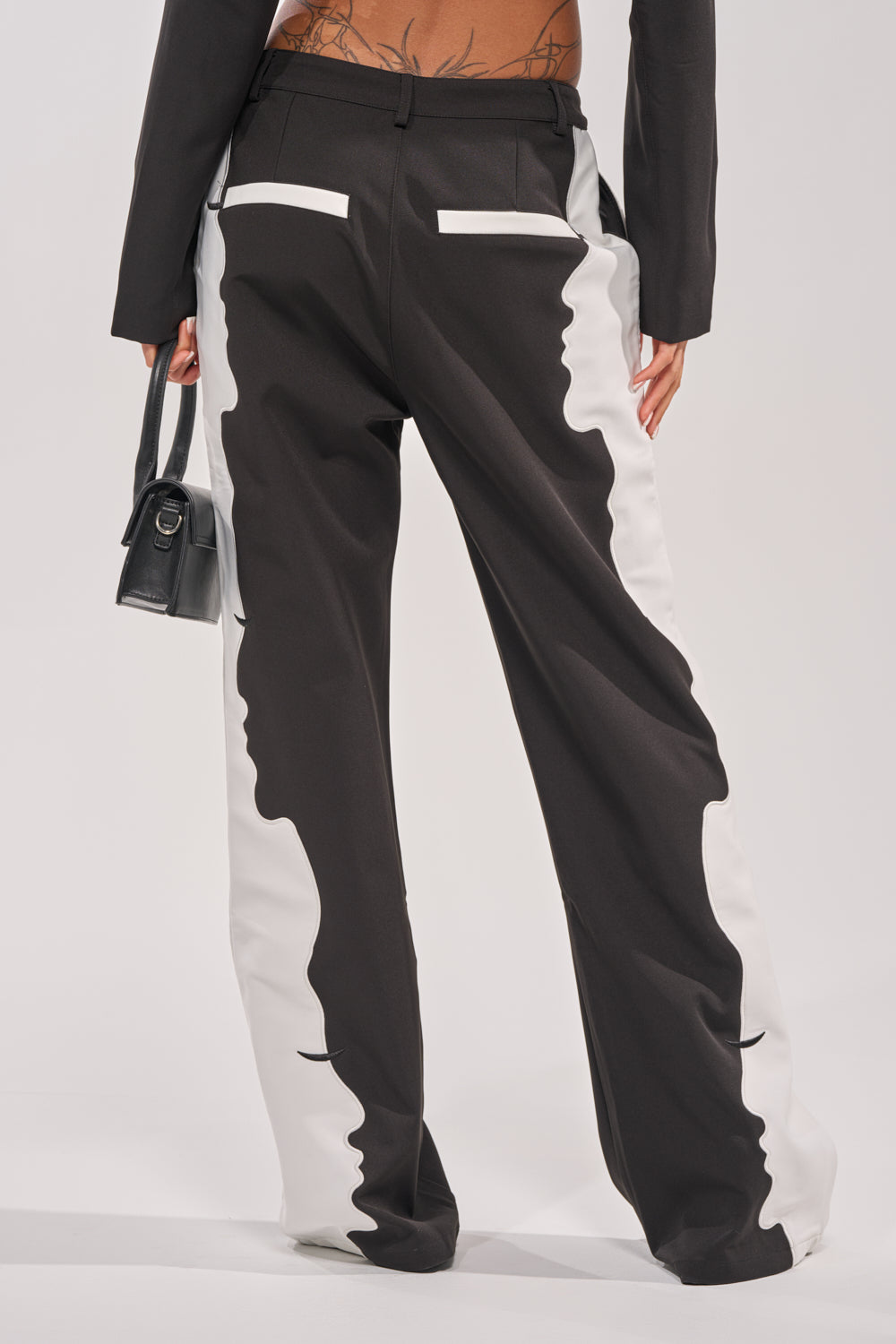 A person is shown from the back wearing POV GRAPHIC TROUSER, featuring a high-rise black and white graphic design with abstract face profiles and a wide-leg silhouette. They have a tattooed lower back, a black jacket, and hold a small black handbag. 4