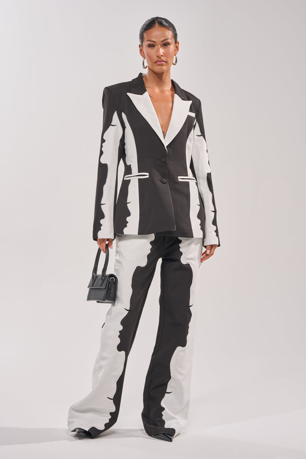 A woman exudes confidence in POV GRAPHIC TROUSER, a bold black-and-white suit with abstract, wavy color-block patterns. She accessorizes with a sleek black handbag and hoop earrings, her hair slicked back for a polished look. 5