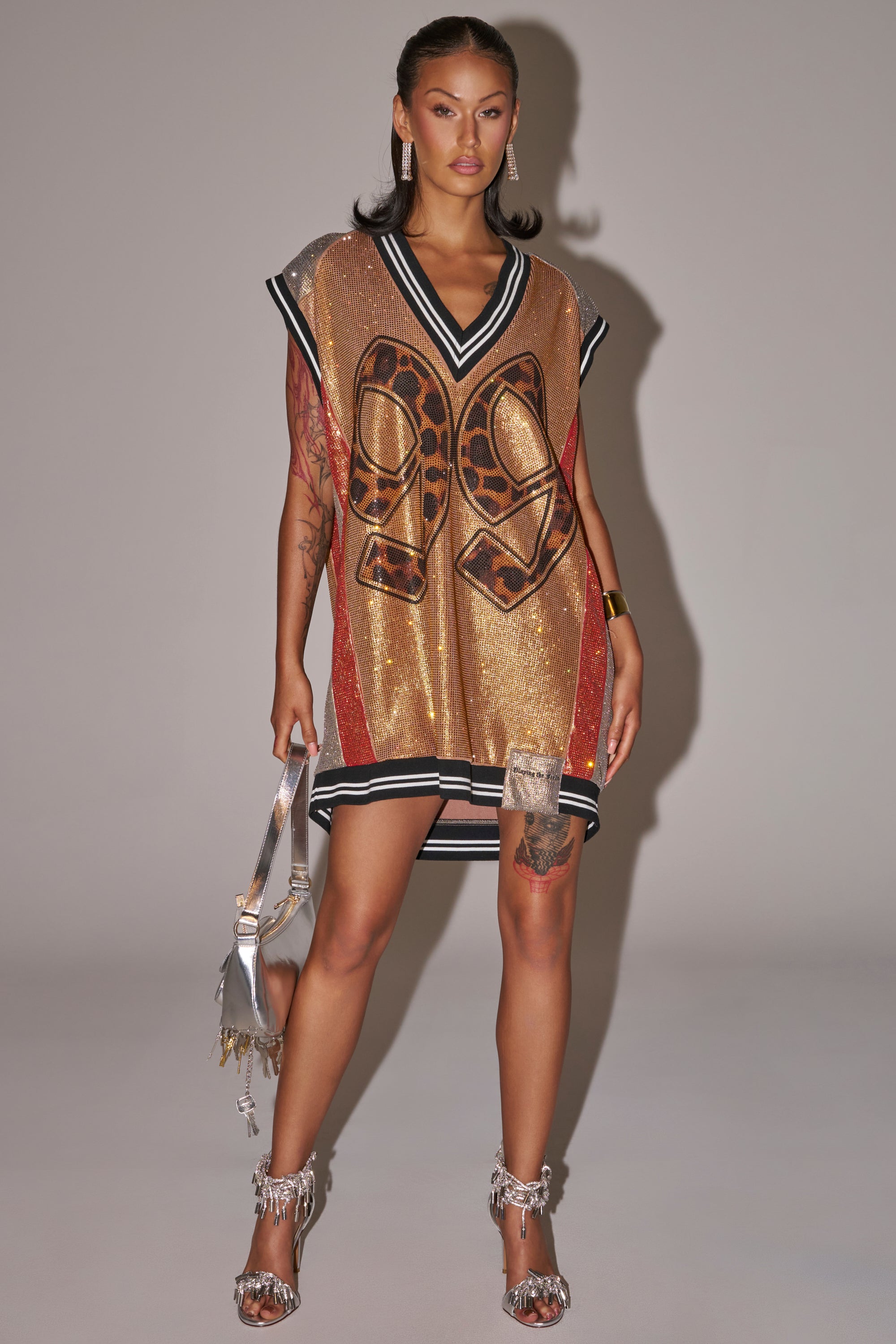 A woman poses against a plain background in the REESE COLORBLOCK RHINESTONE SLEEVELESS JERSEY MINI DRESS with “99” on the front, styled for game day glam with silver fringe heels, a metallic bag, and visible tattoos on her arm and leg. 0
