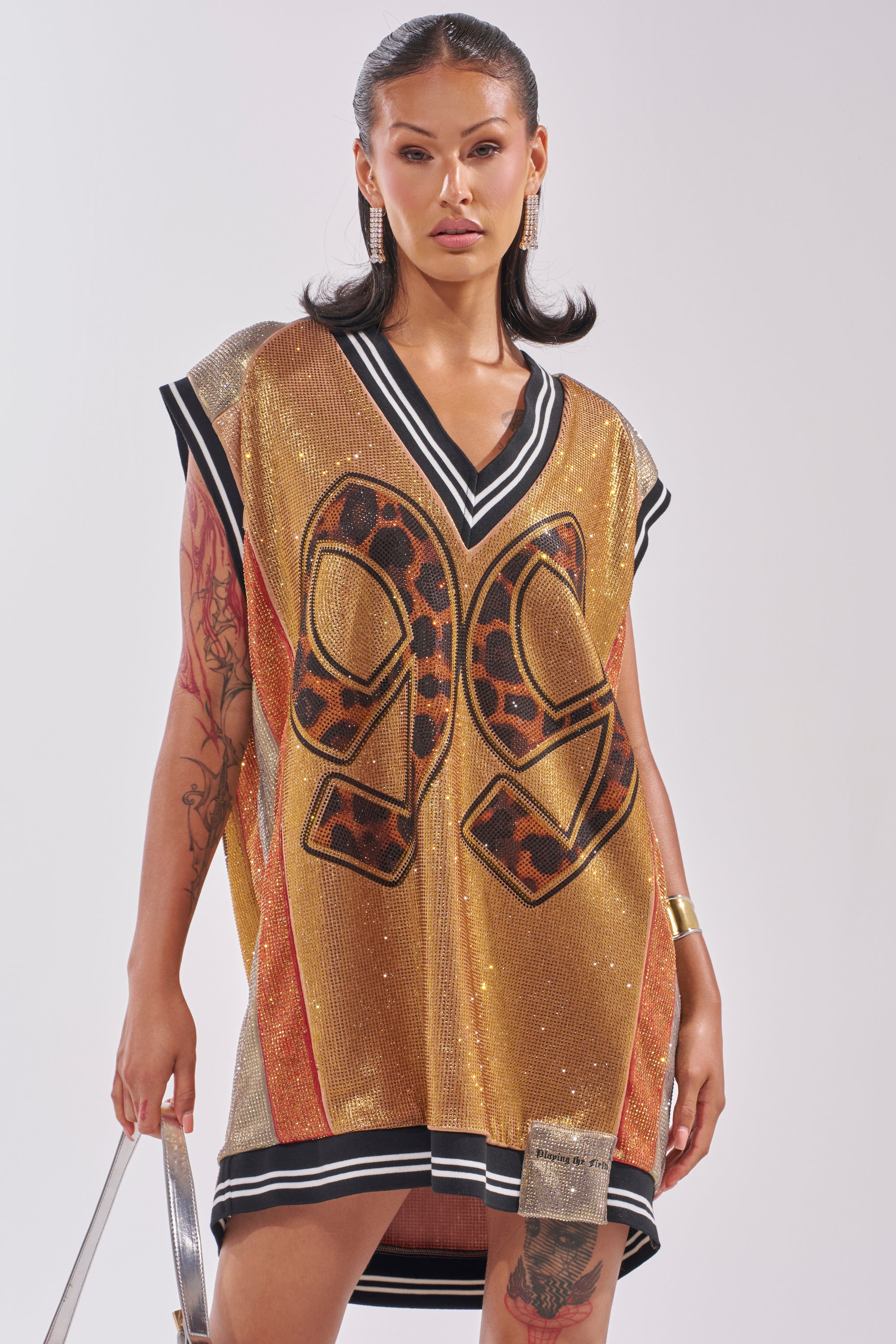 A woman strikes a confident pose in the REESE COLORBLOCK RHINESTONE SLEEVELESS JERSEY MINI DRESS, featuring gold sequins, black and leopard print "96," striped trim, and bold accessories for standout game day glam. 1