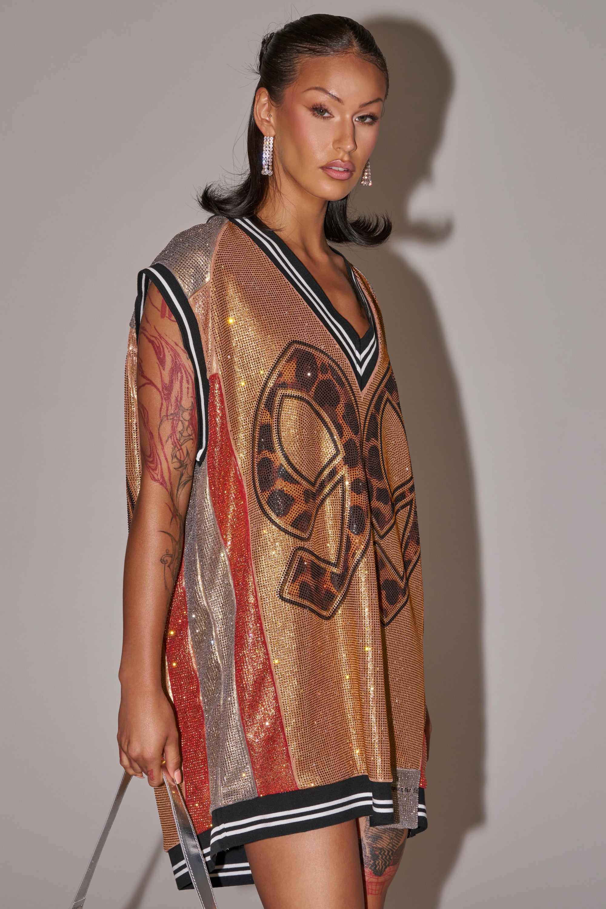 A tattooed woman styles the REESE COLORBLOCK RHINESTONE SLEEVELESS JERSEY MINI DRESS—a gold and orange sequined dress with black and white trim, bold front details, long earrings, and a strap, channeling game day glam against a neutral background. 2