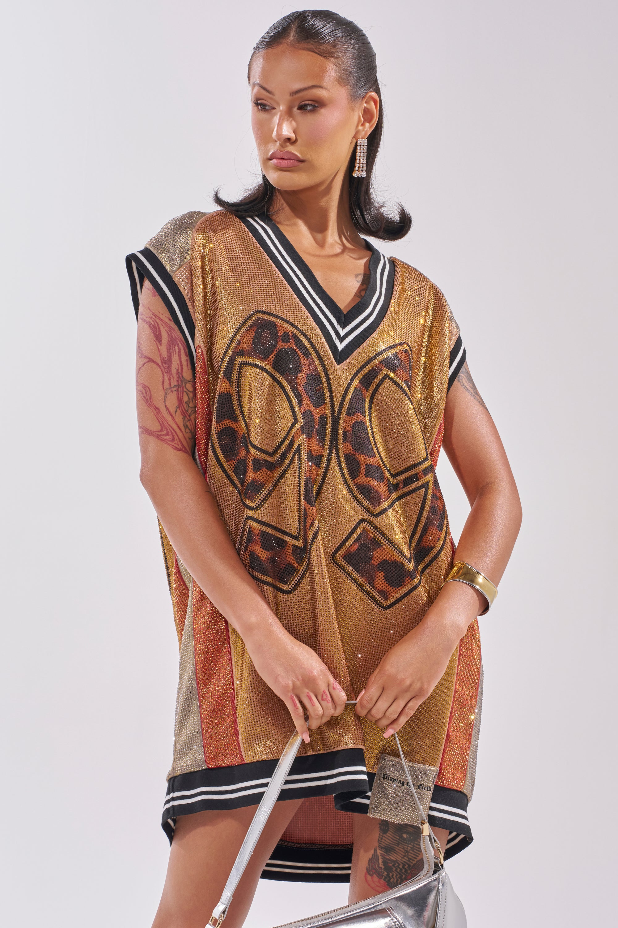 A woman channels courtside glamour in the REESE COLORBLOCK RHINESTONE SLEEVELESS JERSEY MINI DRESS, accessorized with tattoos, hoop earrings, a silver handbag, and slicked-back hair. 3
