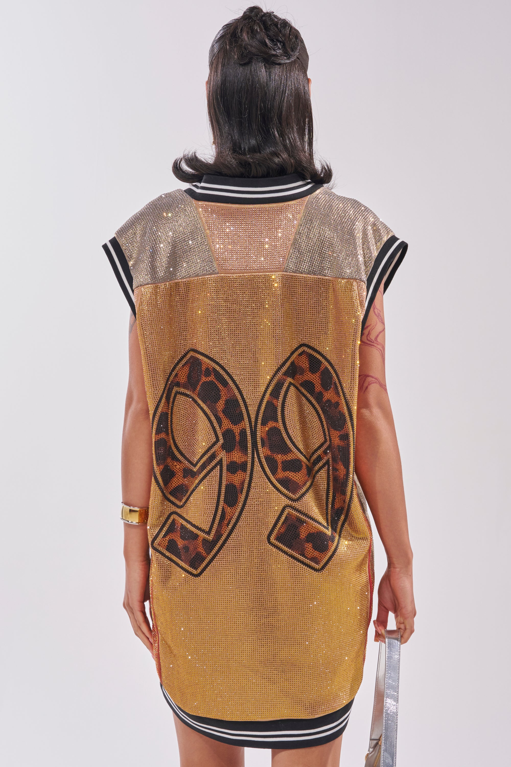 A person faces away from the camera, highlighting courtside chic in the REESE COLORBLOCK RHINESTONE SLEEVELESS JERSEY MINI DRESS with gold and black sequins, a leopard print "99" back, dark hair, and a silver bag for a sparkling finish. 5