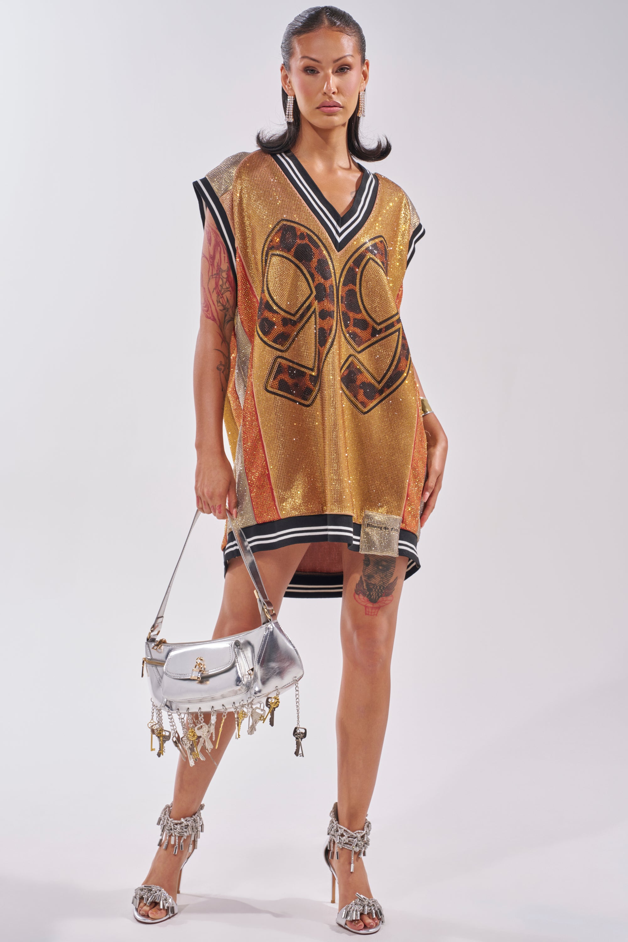 A woman channels courtside glamour in the REESE COLORBLOCK RHINESTONE SLEEVELESS JERSEY MINI DRESS, styled with a silver handbag and high heels, showing off her tattoos and slicked-back hair. 6