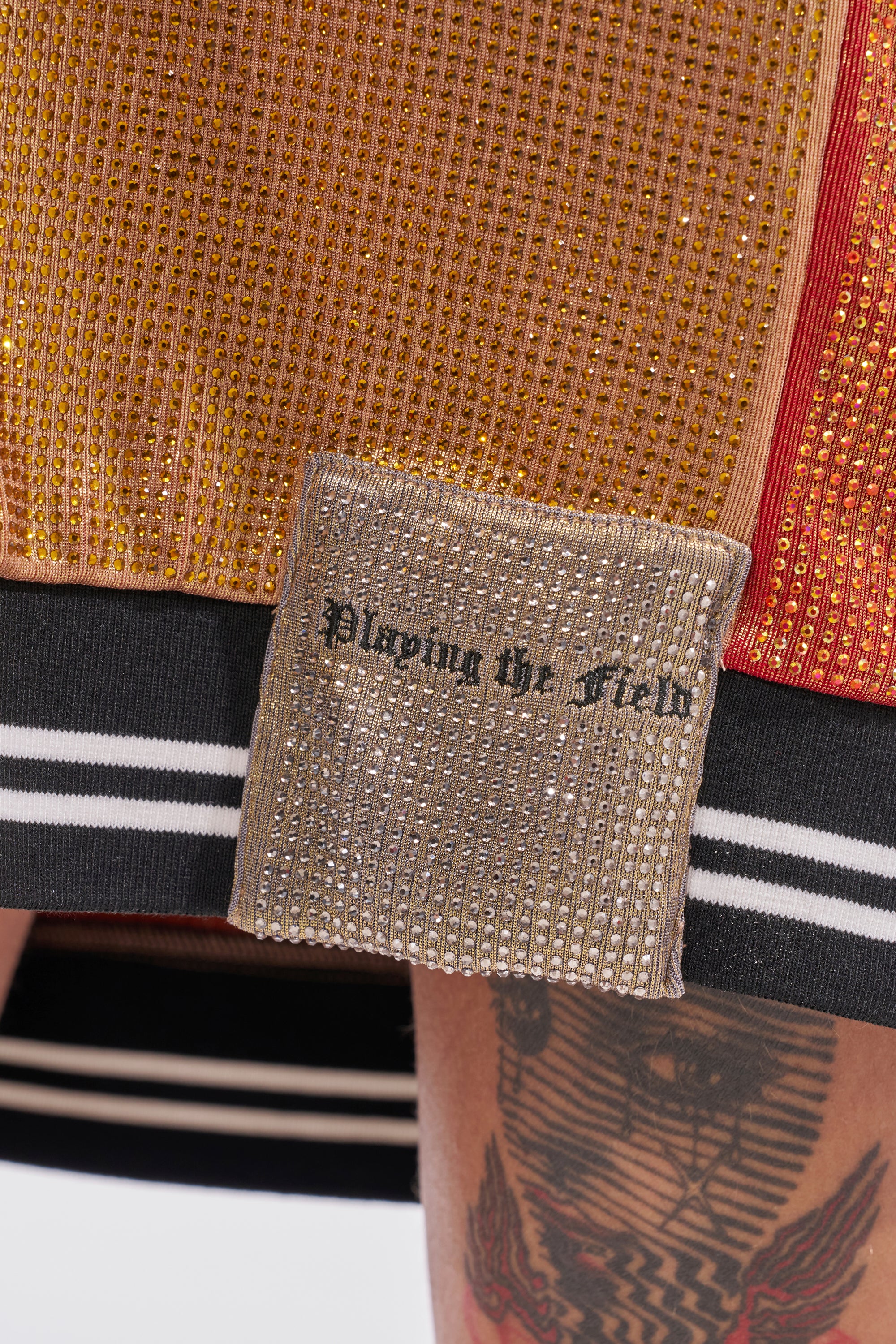 A close-up of a person in the REESE COLORBLOCK RHINESTONE SLEEVELESS JERSEY MINI DRESS, featuring gold and orange textures, a metallic "Playing the Field" patch, black and white striped trim, and part of a tattooed leg. 7
