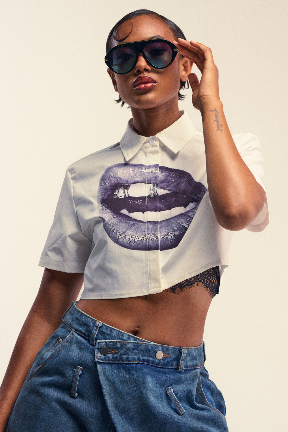 A person wearing sunglasses and the ICE ME OUT BUTTON DOWN TOP, featuring a striking white lip graphic and cropped button-down design, pairs it with loose blue jeans and poses confidently with one hand adjusting their glasses, showcasing bold streetwear style.