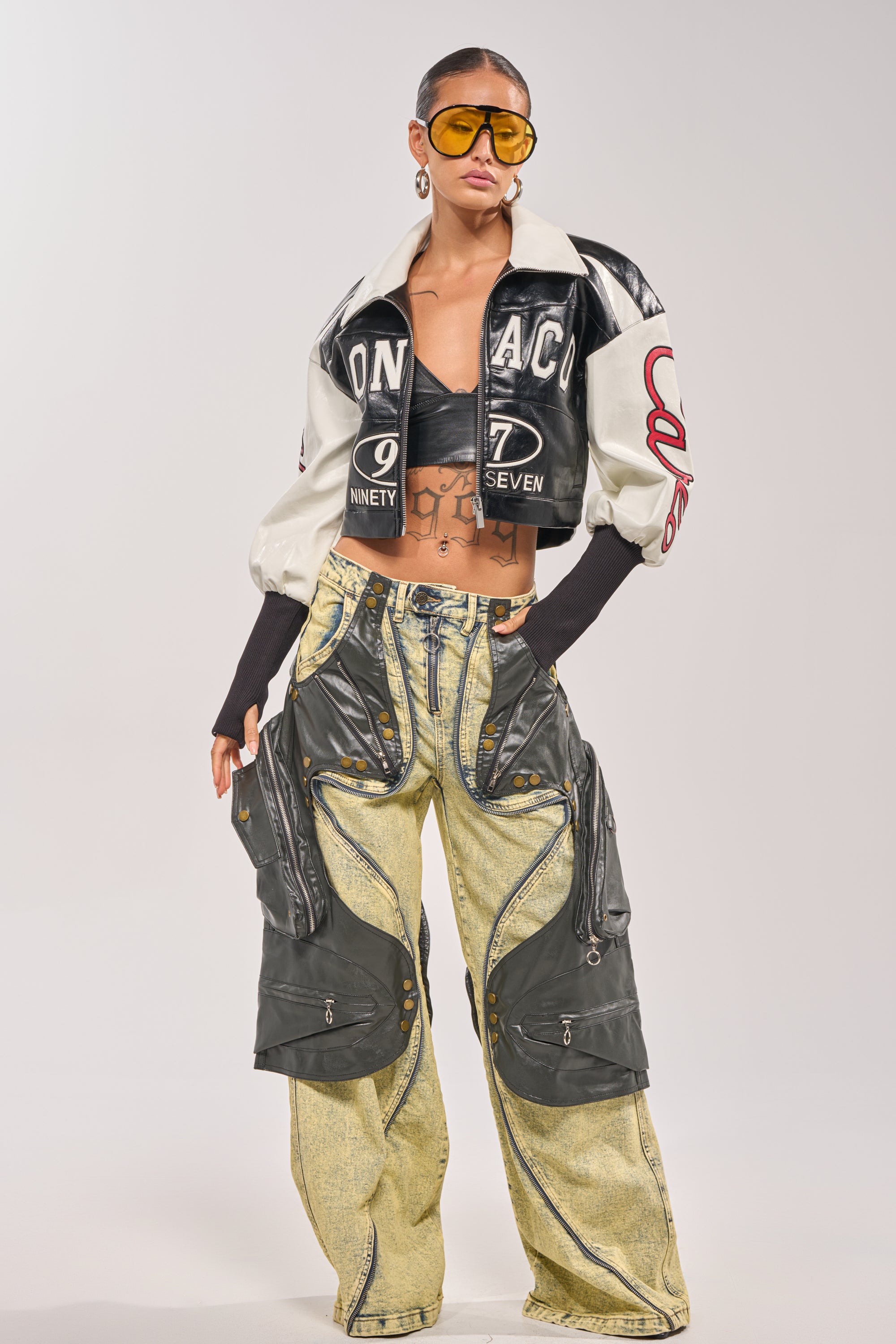 A woman poses in a bold look featuring the LET'S RACE EMBROIDERED BOMBER, a black and white cropped jacket, paired with a black bralette, yellow cargo pants with zippers, oversized yellow sunglasses, and large hoop earrings. 8