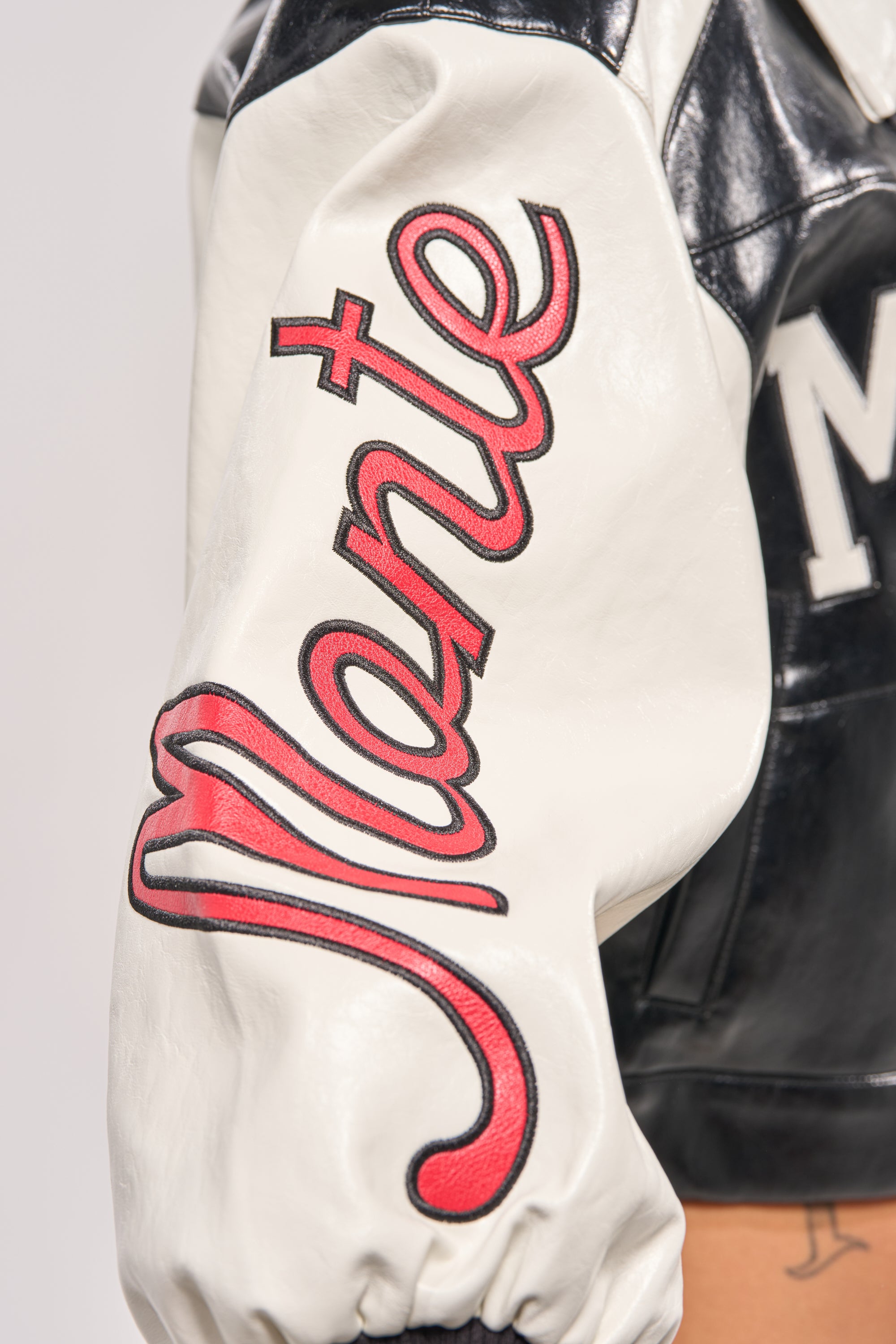 Close-up of the LET'S RACE EMBROIDERED BOMBER's black and white sleeve, showcasing "Monte" in bold red cursive with black outline, plus black leather detailing on the cropped bomber jacket’s shoulder. 10