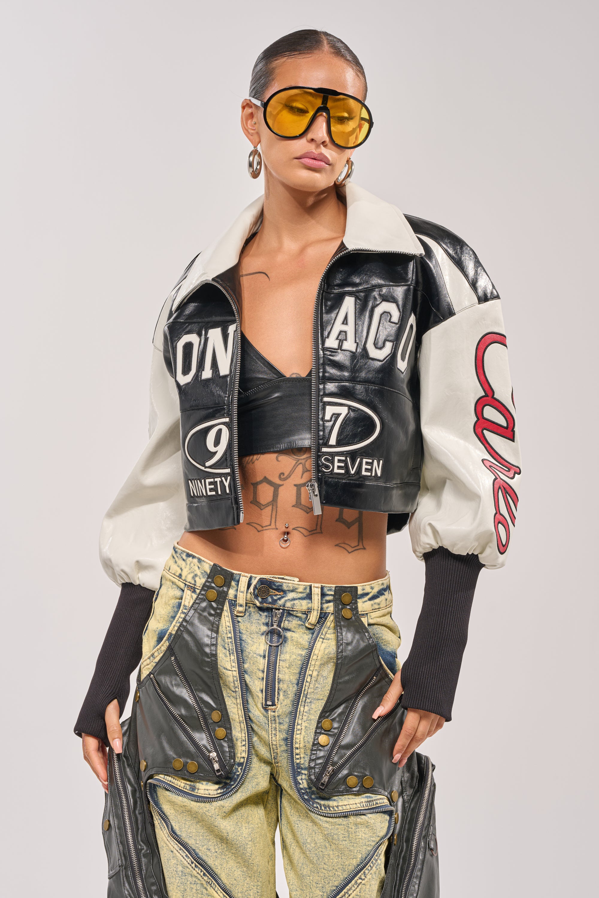 A person poses confidently in the LET'S RACE EMBROIDERED BOMBER, oversized yellow sunglasses, large hoop earrings, and distinctive high-waisted pants with zipper and leather details. 2