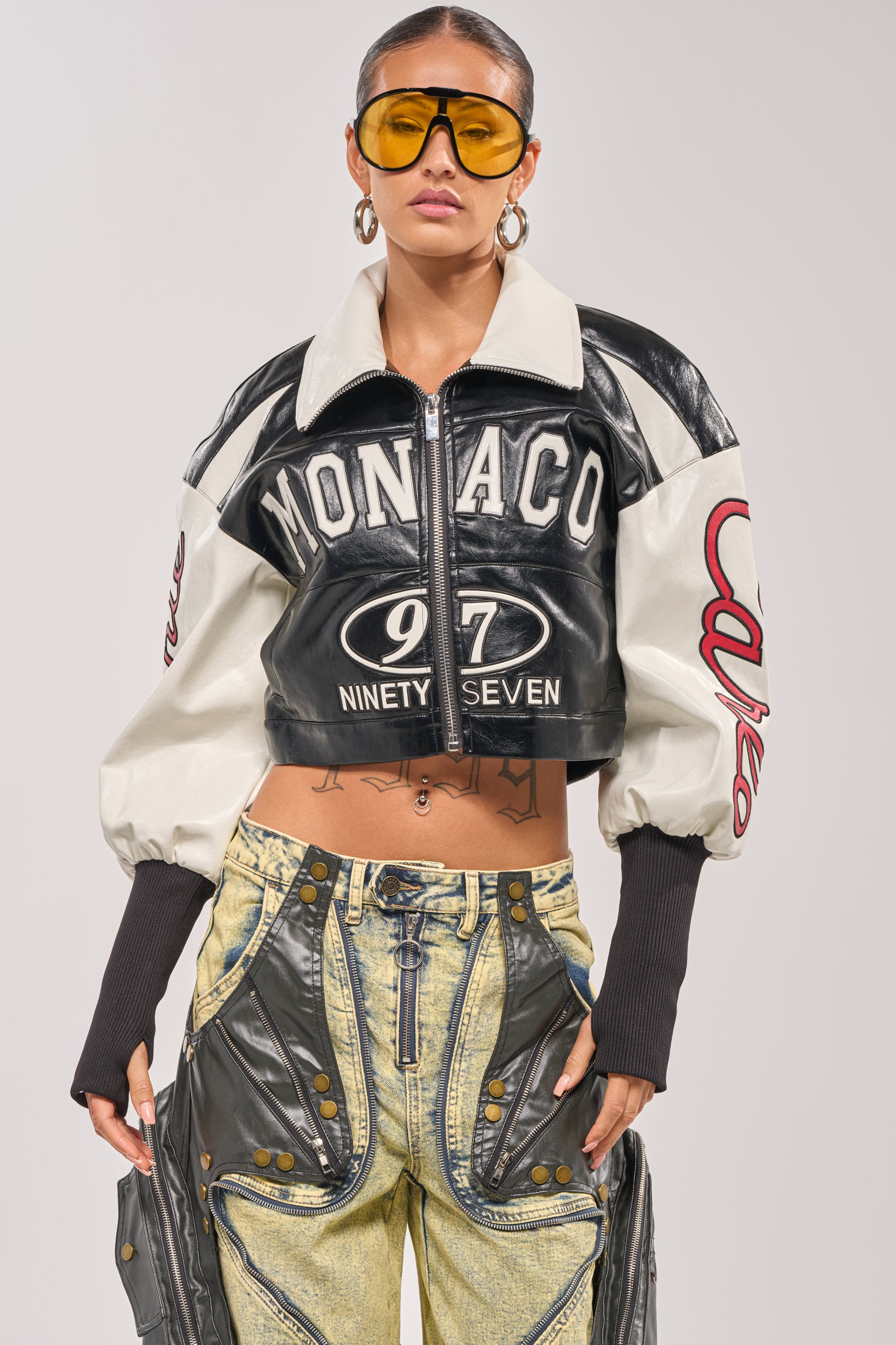 Someone poses against a plain background in oversized yellow sunglasses, baggy denim pants with button and stitch details, and the LET'S RACE EMBROIDERED BOMBER—a cropped black and white jacket with "MONACO 97 NINETY SEVEN" on the front. 3