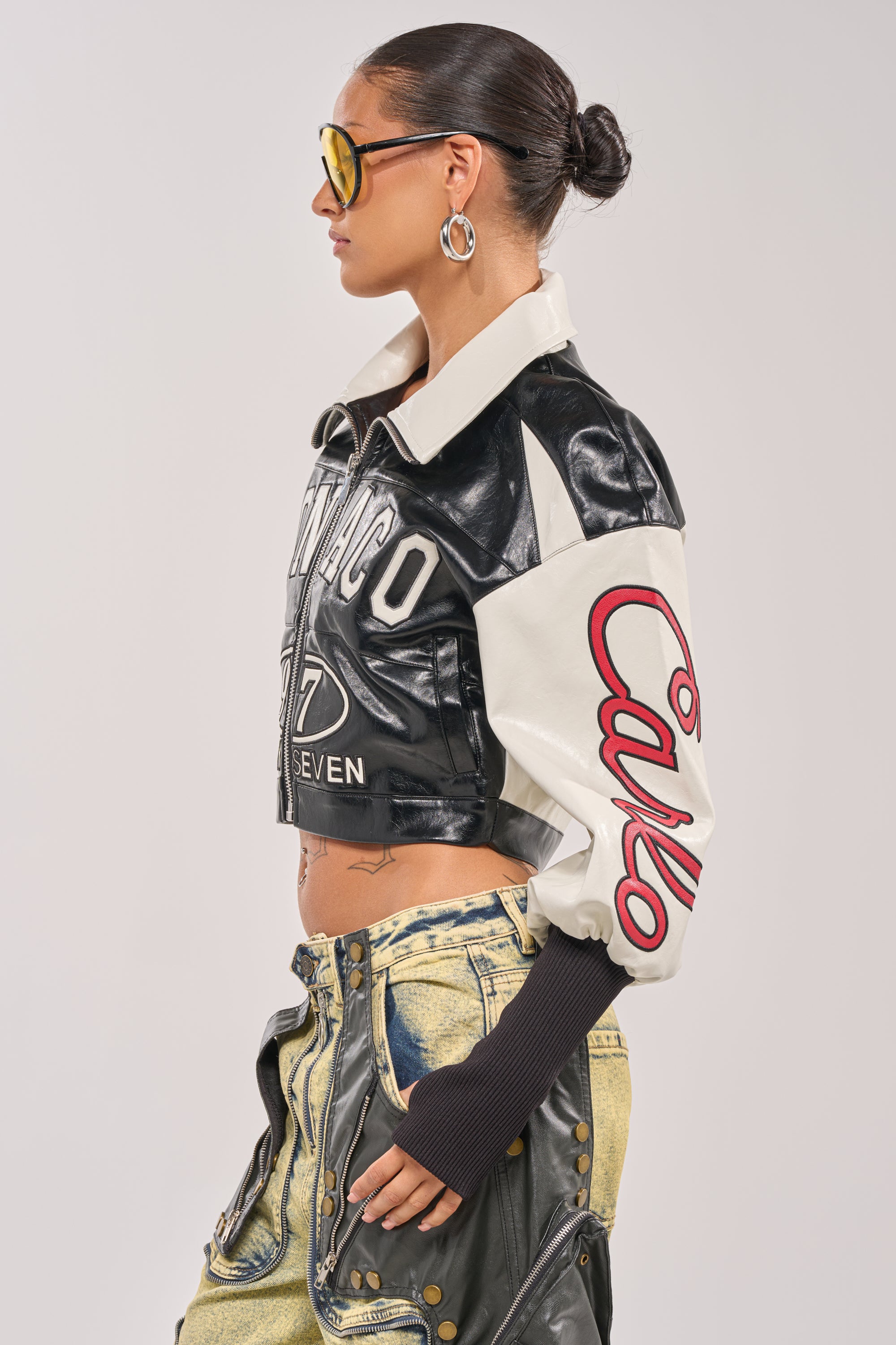 A person with slicked-back hair, large hoop earrings, and yellow sunglasses wears the LET'S RACE EMBROIDERED BOMBER, a black and white cropped jacket with bold designs, paired with low-rise yellow jeans. 5