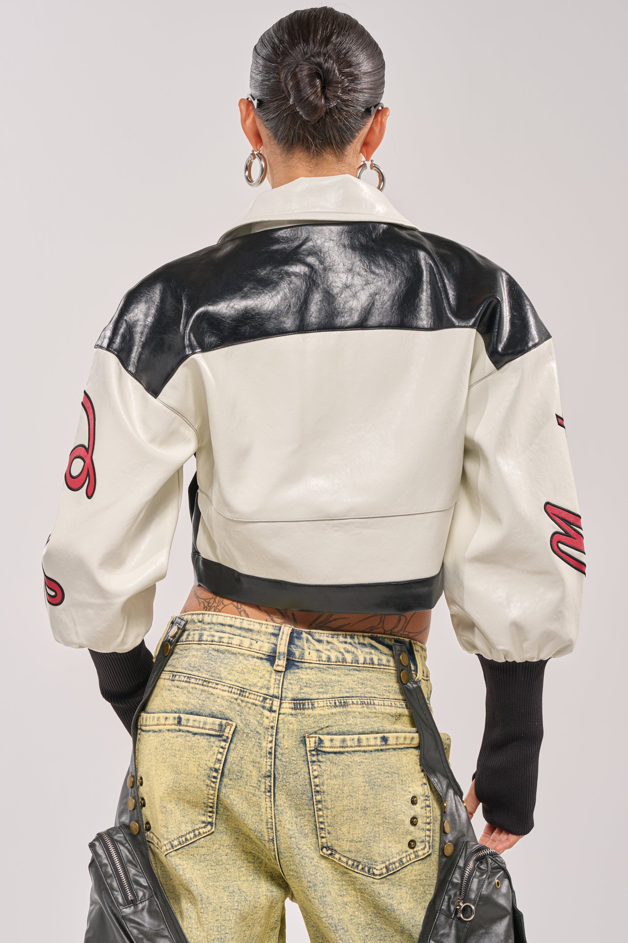 Seen from the back, a person with slicked-back hair wears the LET'S RACE EMBROIDERED BOMBER—black and white with red sleeve patches—paired with high-waisted, acid-wash yellow jeans. 6