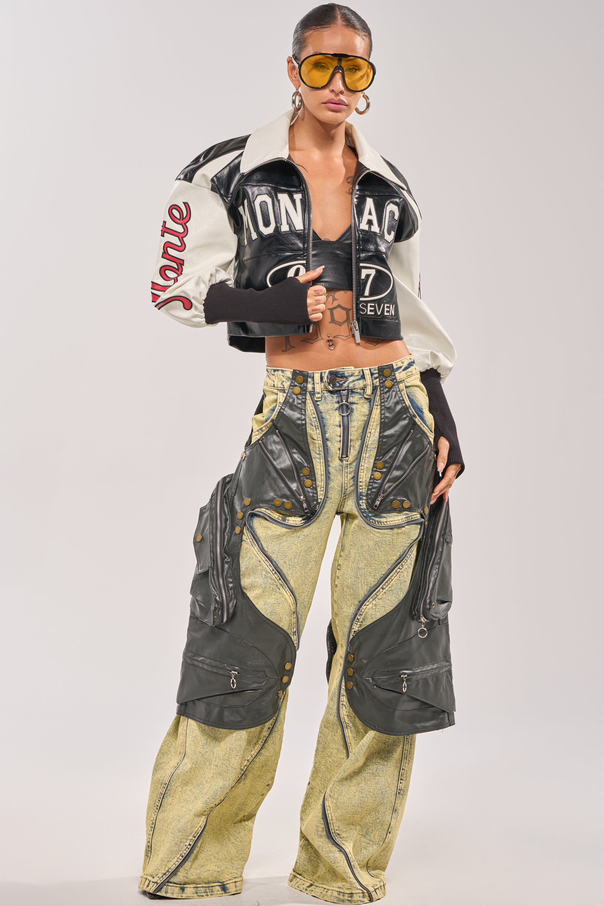 A woman poses confidently in the LET'S RACE EMBROIDERED BOMBER, styled with oversized yellow aviator sunglasses and wide-leg yellow denim pants featuring black faux leather details and zipper accents. 7