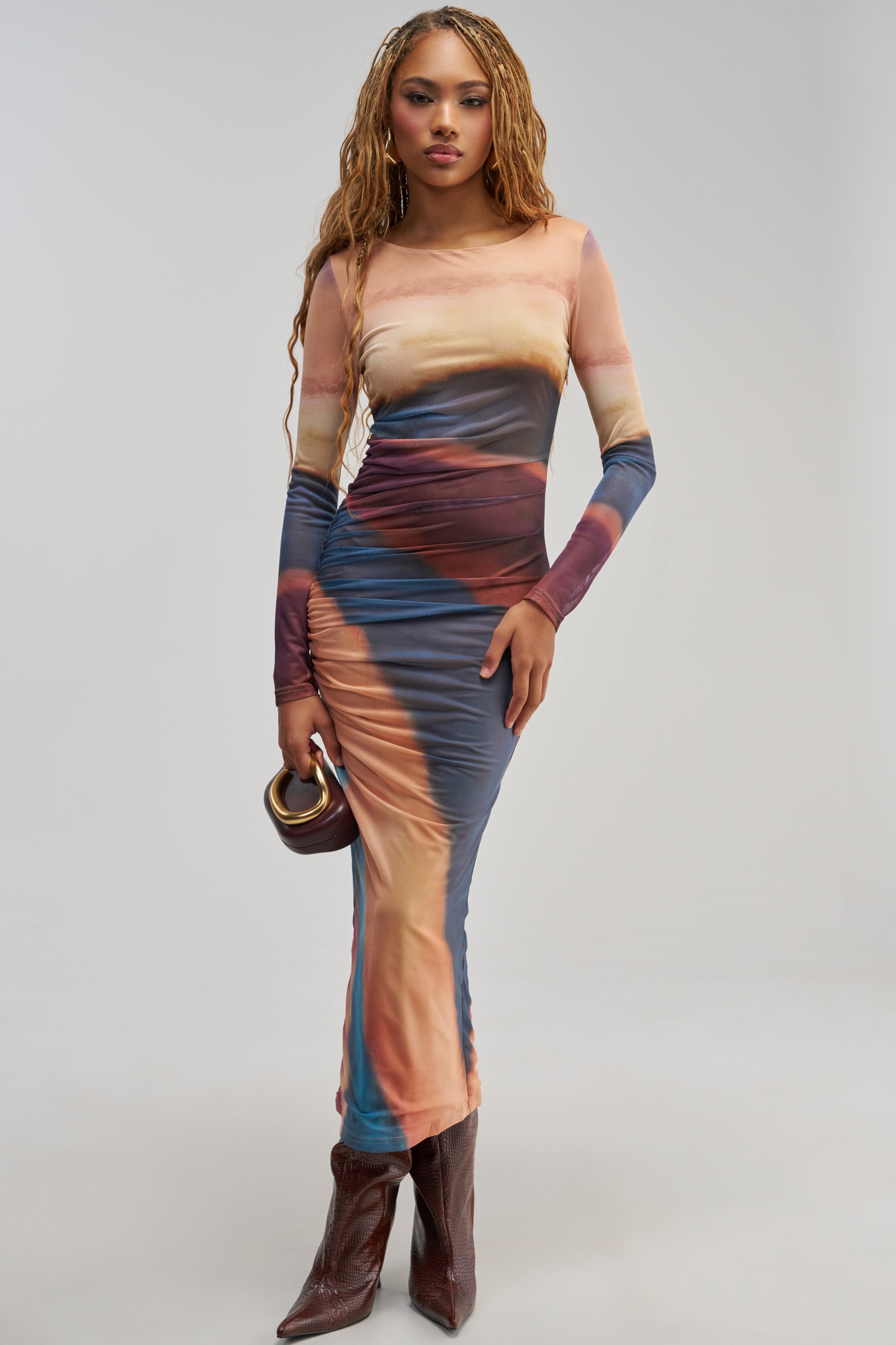 Wearing the WILD GLOW PRINTED MESH MAXI DRESS—a long-sleeve, multicolored fitted evening dress featuring abstract patterns and ruched detailing—a woman stands against a plain light background, accessorized with a small brown handbag and brown boots.
