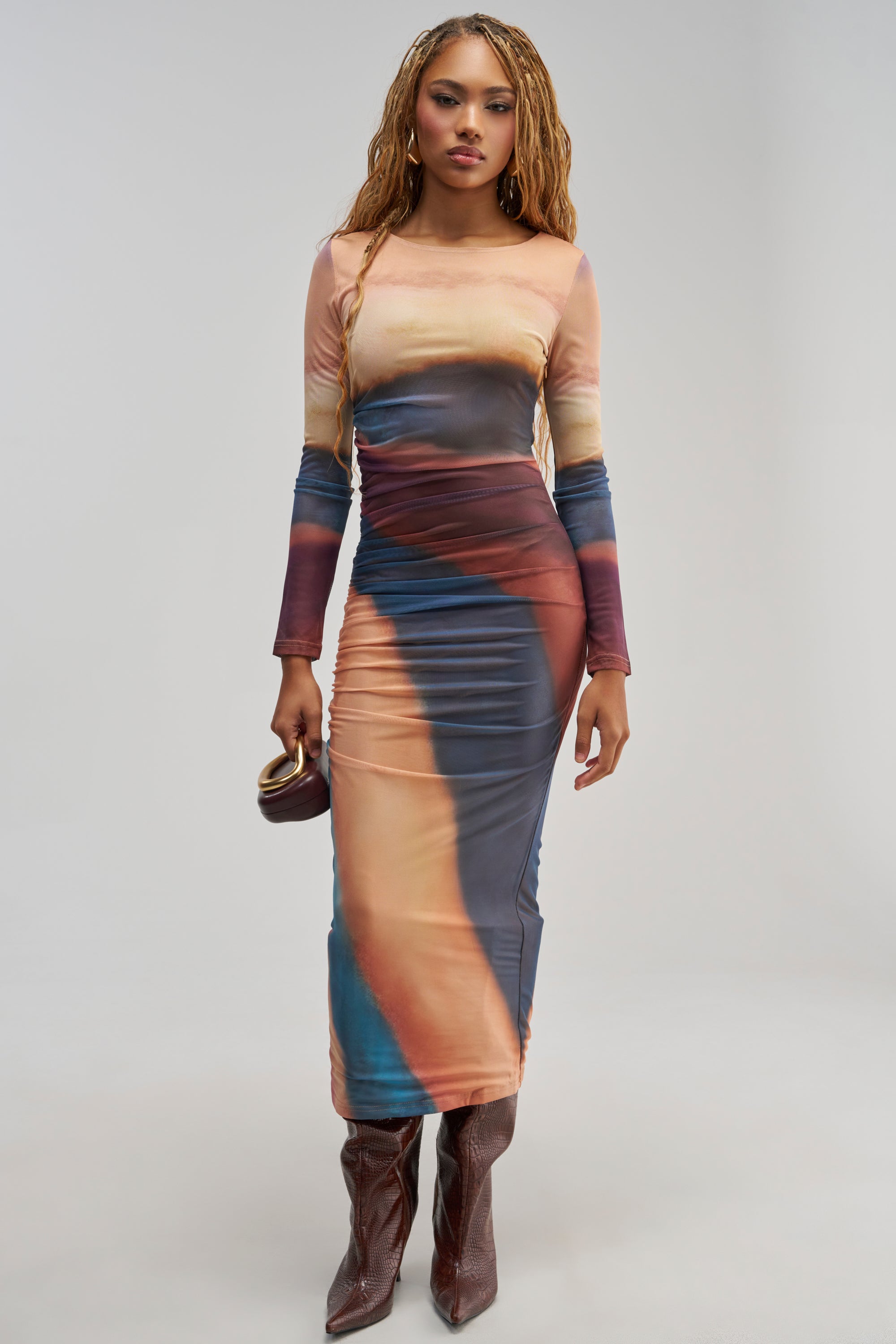 A woman with long braided hair wears the WILD GLOW PRINTED MESH MAXI DRESS, a long-sleeved, fitted ombre mesh maxi dress featuring abstract multicolor patterns. She pairs it with brown boots, holds a small clutch, and stands against a light grey background.