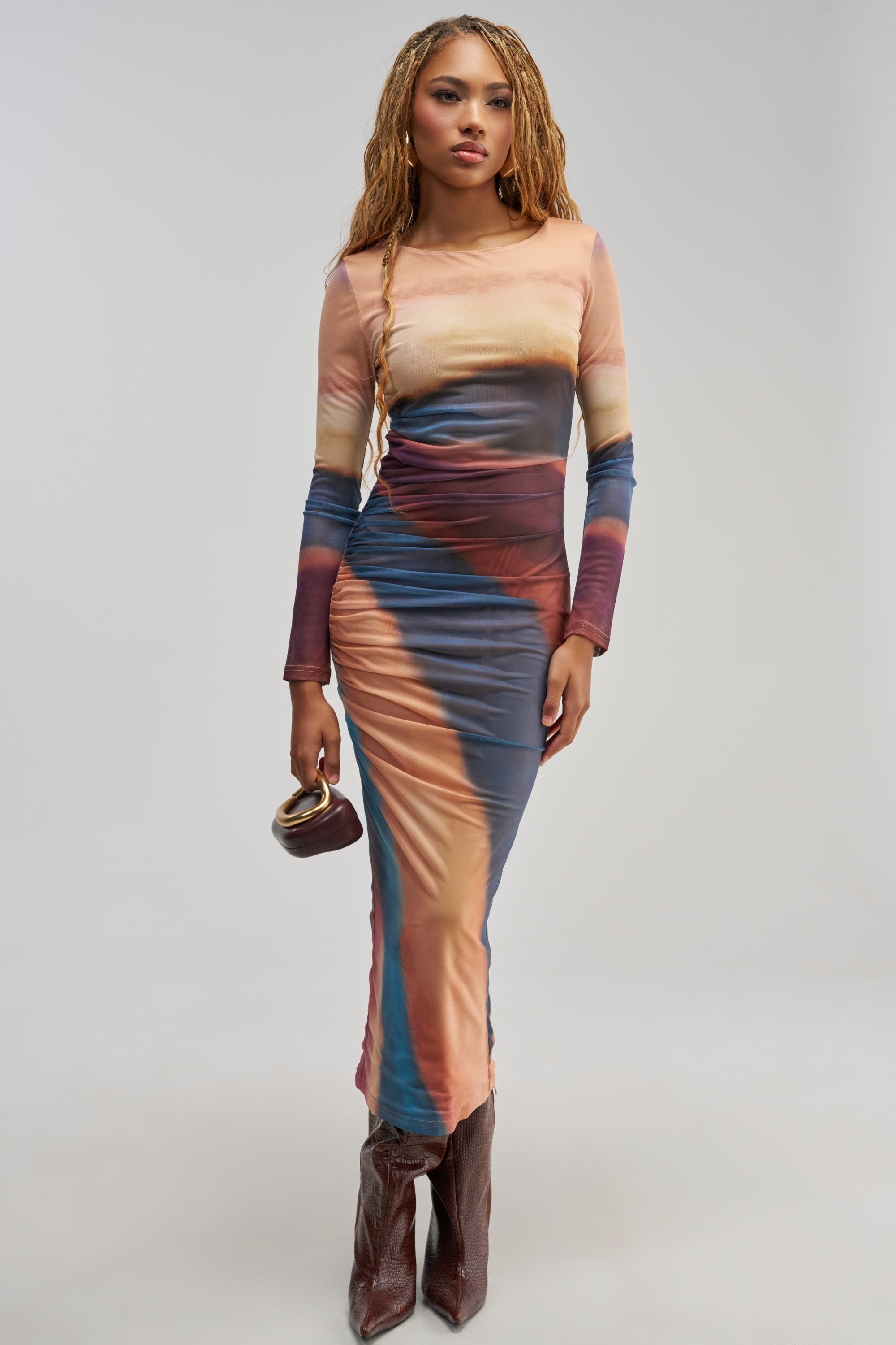 A woman with long braids wears the WILD GLOW PRINTED MESH MAXI DRESS—a colorful, abstract-patterned, ombre mesh maxi dress with long sleeves and ruched detailing—paired with brown boots and a small brown purse against a plain background.