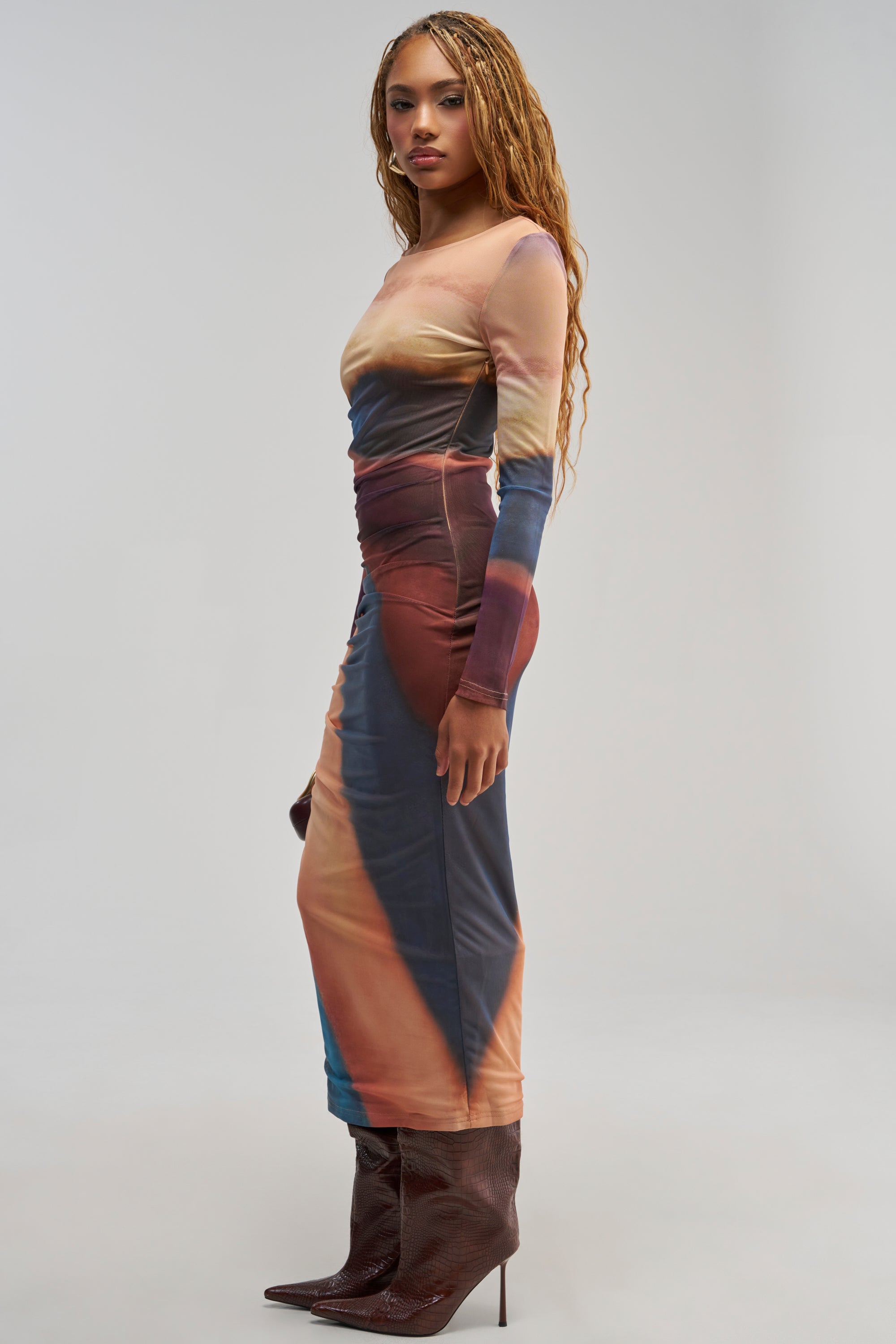 A woman with long braids stands in profile against a plain light gray background, wearing the WILD GLOW PRINTED MESH MAXI DRESS—a fitted, long-sleeve ombre mesh maxi dress with ruched detailing—paired with pointed brown boots.