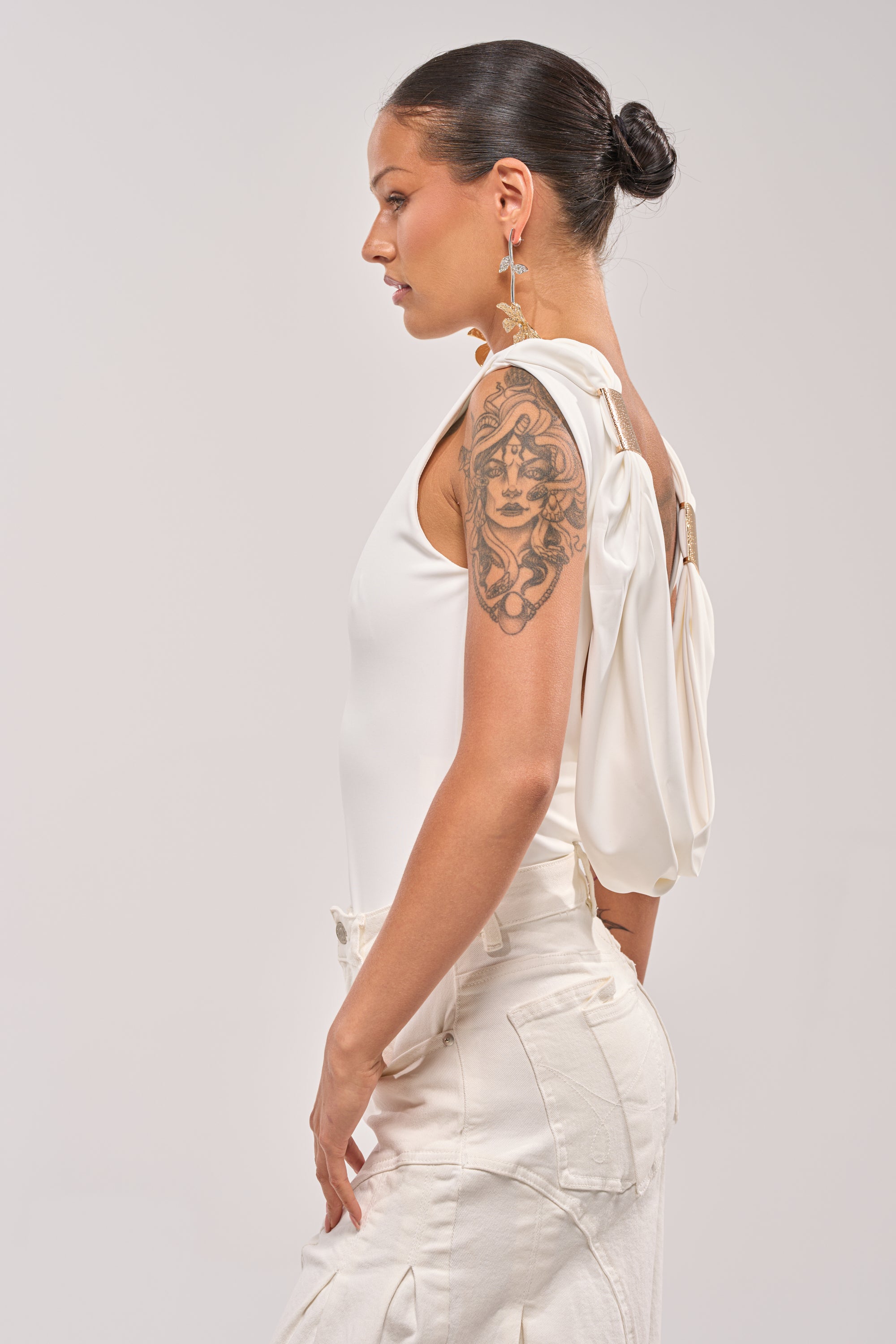 A woman with a Medusa tattoo on her upper arm wears the STAY CLASSY STRETCH BODYSUIT, featuring draped fabric on one shoulder and styled with high-waisted white pants. Her hair is in a sleek bun, and she stands in profile wearing dangling earrings. 2