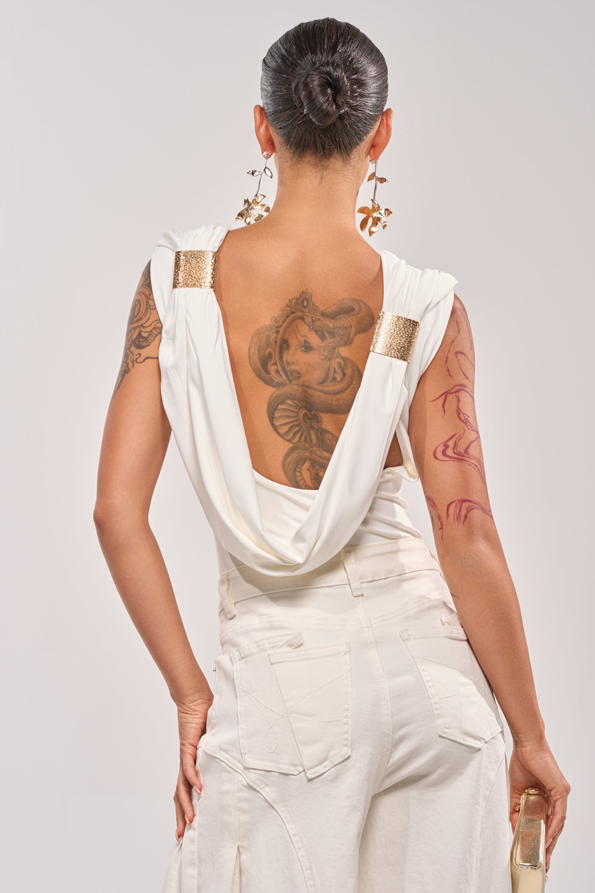 A person with a tattooed back and arm faces away, wearing the STAY CLASSY STRETCH BODYSUIT in white with matching pants. Their hair is in a sleek bun, bold earrings dangle from their ears, and they hold a small gold clutch. 3