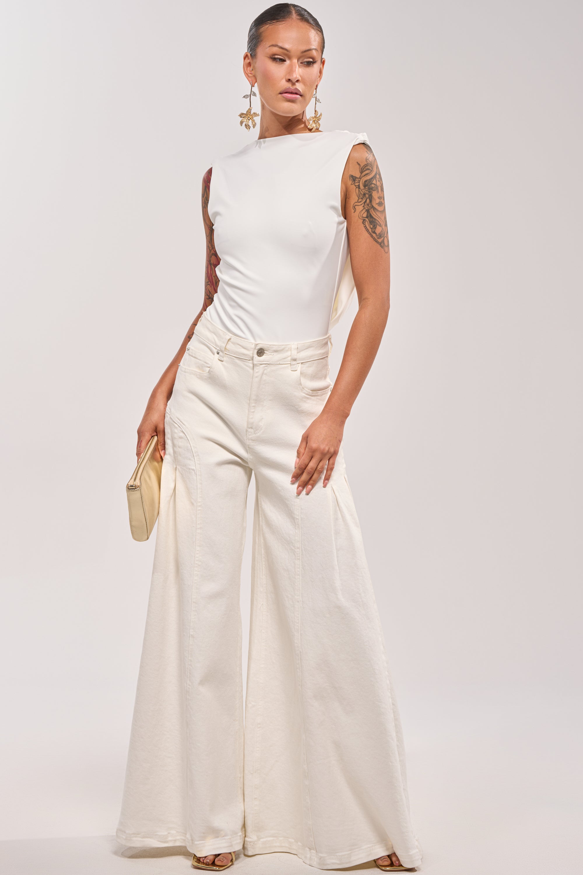 A woman poses confidently in the STAY CLASSY STRETCH BODYSUIT, paired with wide-leg high-waisted pants and statement earrings. She holds a beige clutch, showing off arm tattoos against a light background. 4
