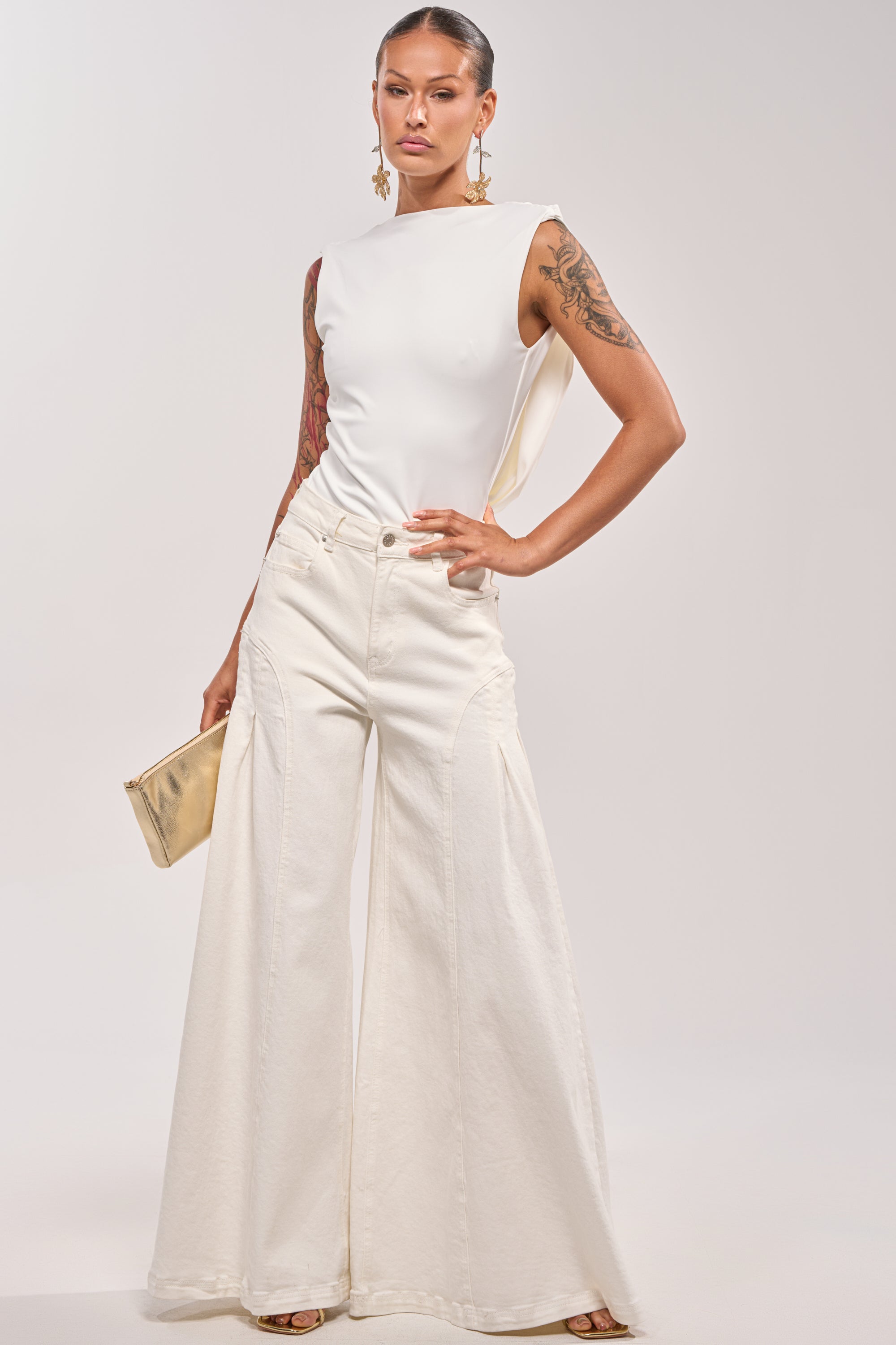 A woman poses confidently in the STAY CLASSY STRETCH BODYSUIT and wide-leg white jeans, holding a small gold clutch. She has arm tattoos, large statement earrings, and her hair is pulled back. 5