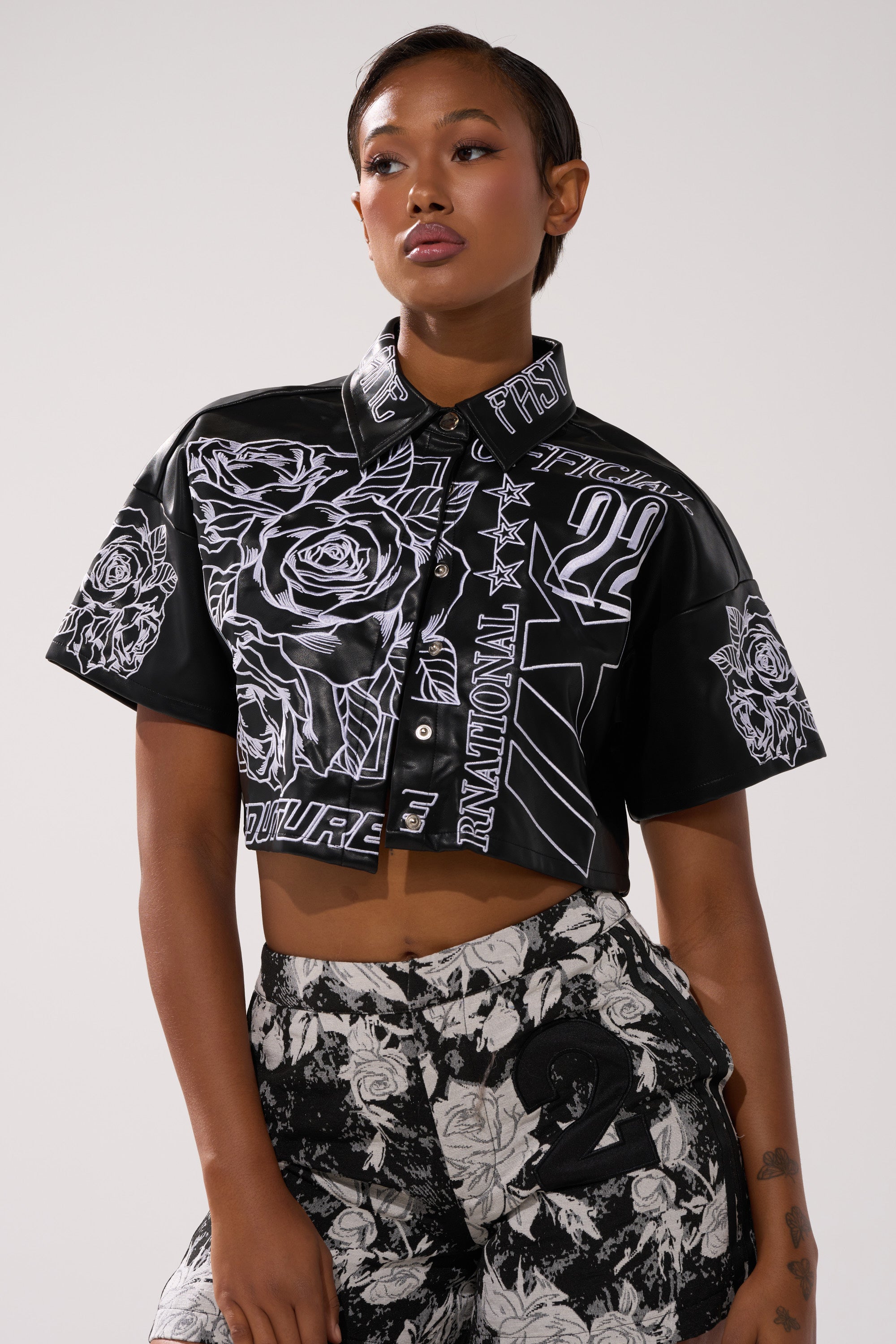 A woman poses confidently in the MAKE IT OFFICIAL FAUX LEATHER BUTTON DOWN IN BLACK, styled with high-waisted shorts and slicked-back hair, standing against a plain background.
