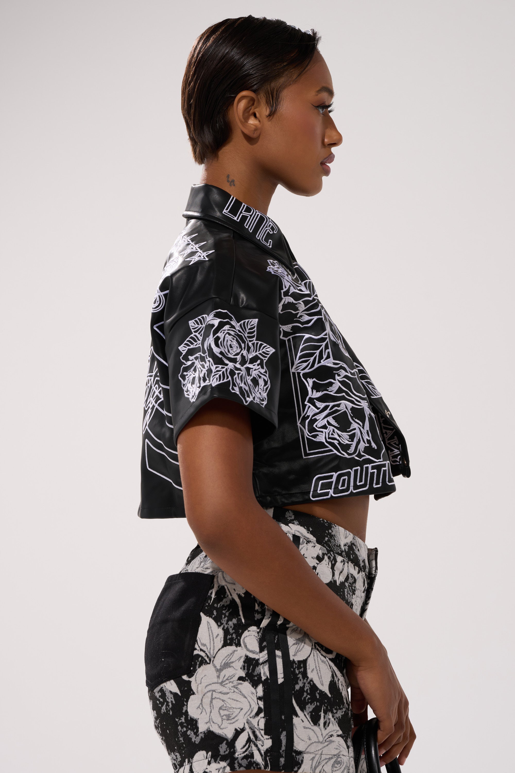 A woman stands in profile against a plain background, wearing the MAKE IT OFFICIAL FAUX LEATHER BUTTON DOWN IN BLACK with matching high-waisted pants. Her hair is slicked back and she holds a small black bag.