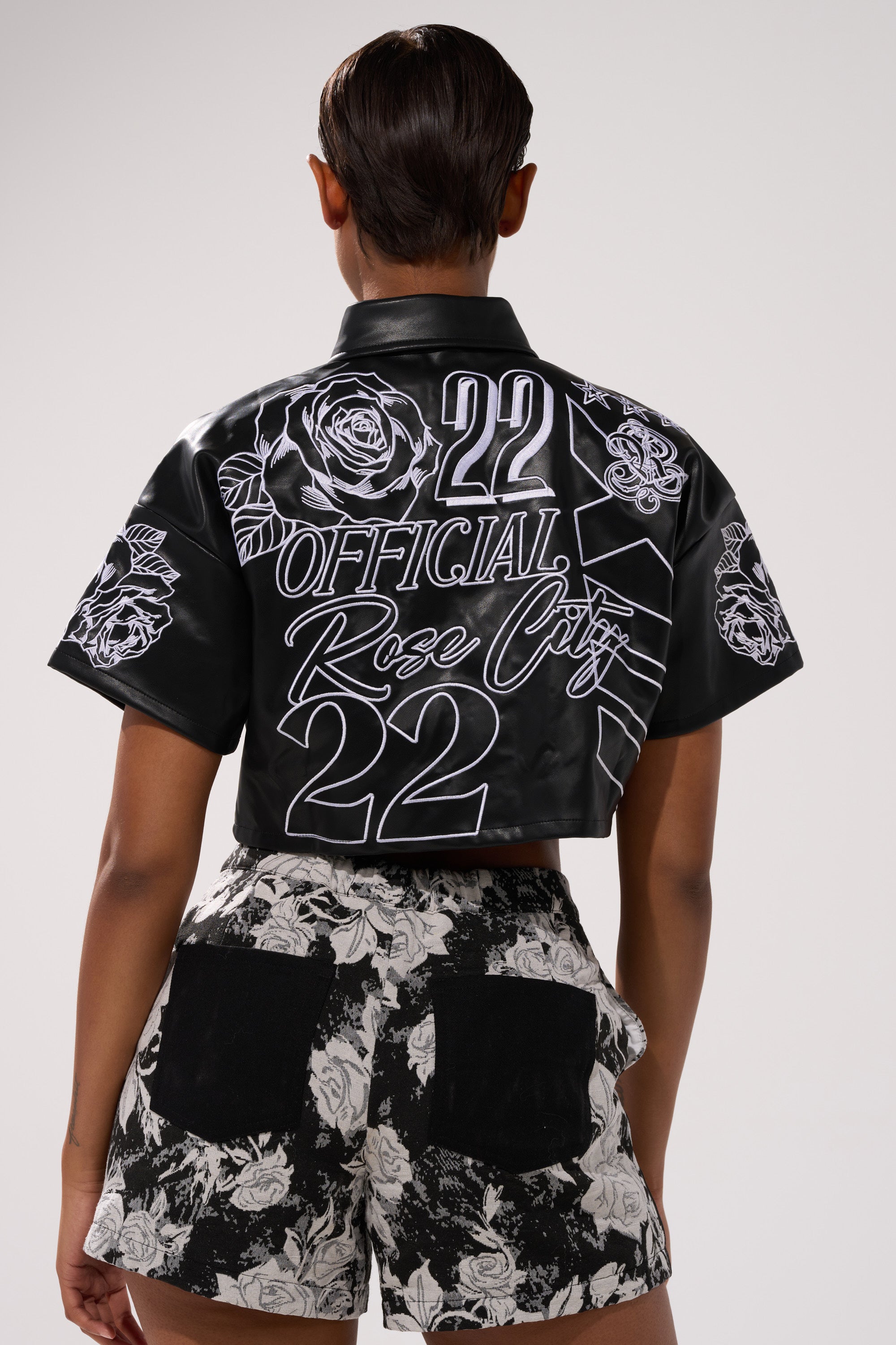 A person facing away wears the MAKE IT OFFICIAL FAUX LEATHER BUTTON DOWN IN BLACK, featuring white graphic designs and "22 OFFICIAL Rose City" text, paired with black-and-white floral shorts with black pockets.